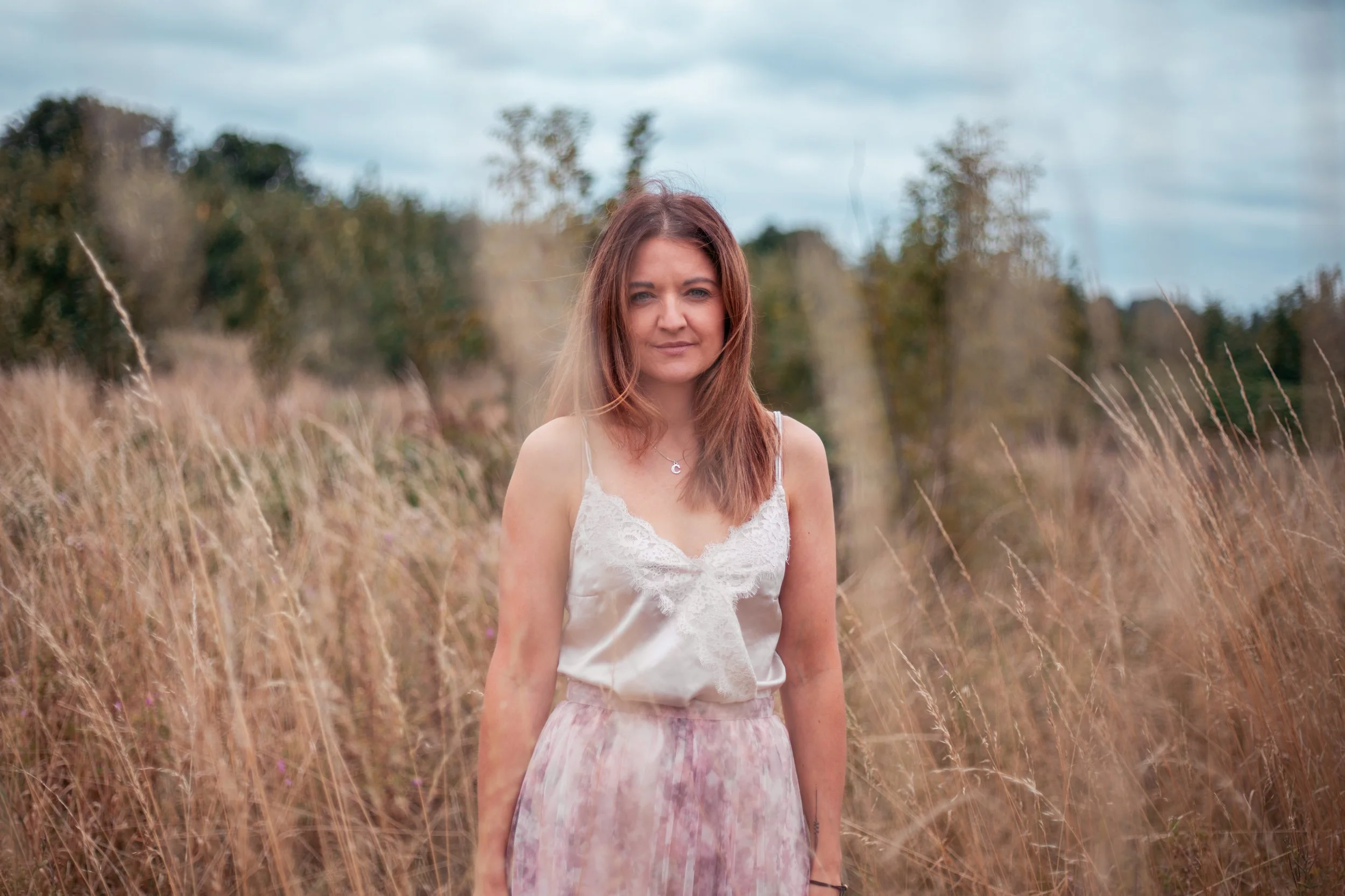 A woman, wearing a white lace top and a flowy pink skirt, standing in a field of tall grasses with trees and cloudy sky in the background. Natural beautiful portrait.