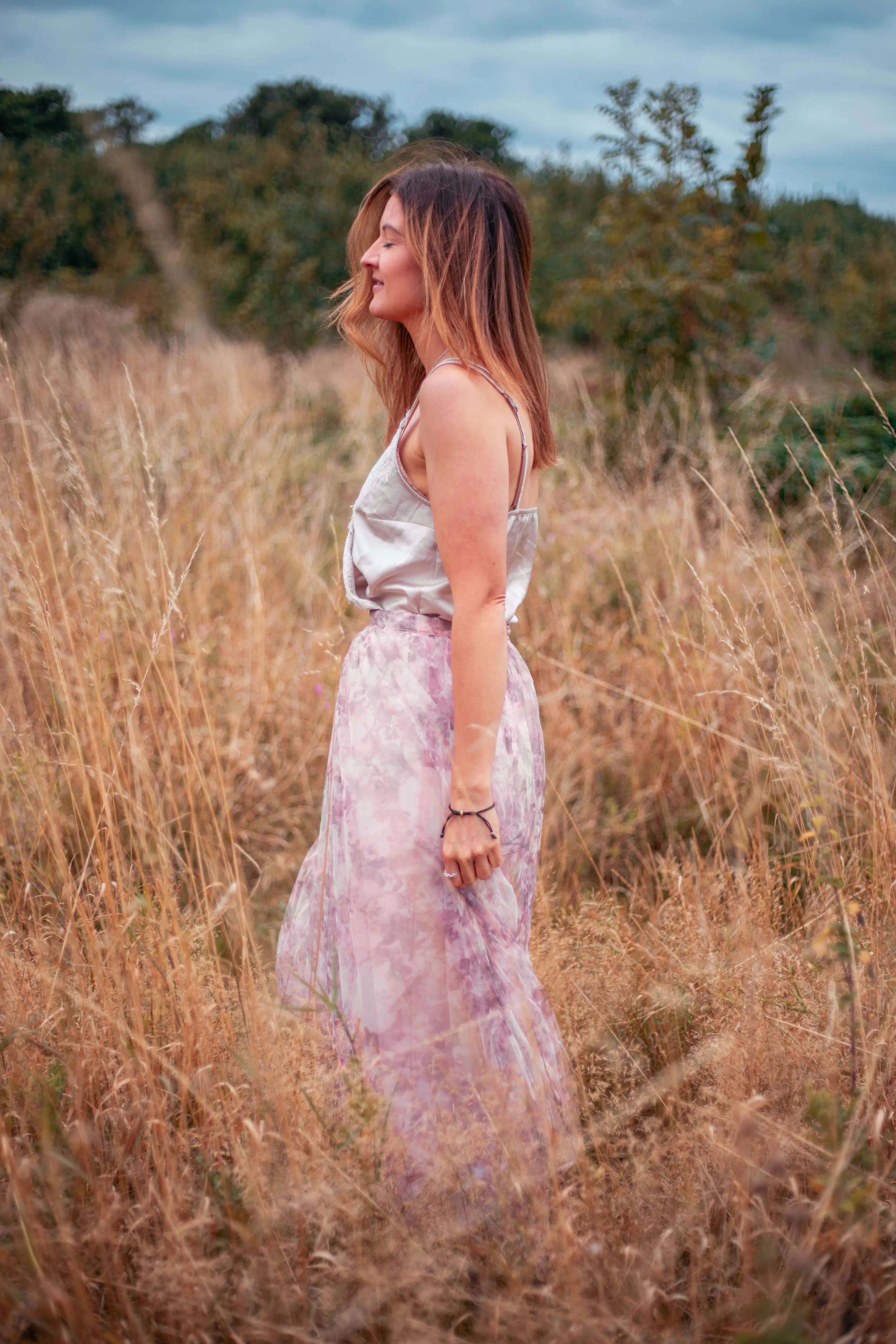 A woman standing in a field of tall, dry grass, smiling with eyes closed, wearing a satin camisole top and a flowy pink tie-dye skirt, with trees and cloudy sky in the background.  Natural beautiful portrait.