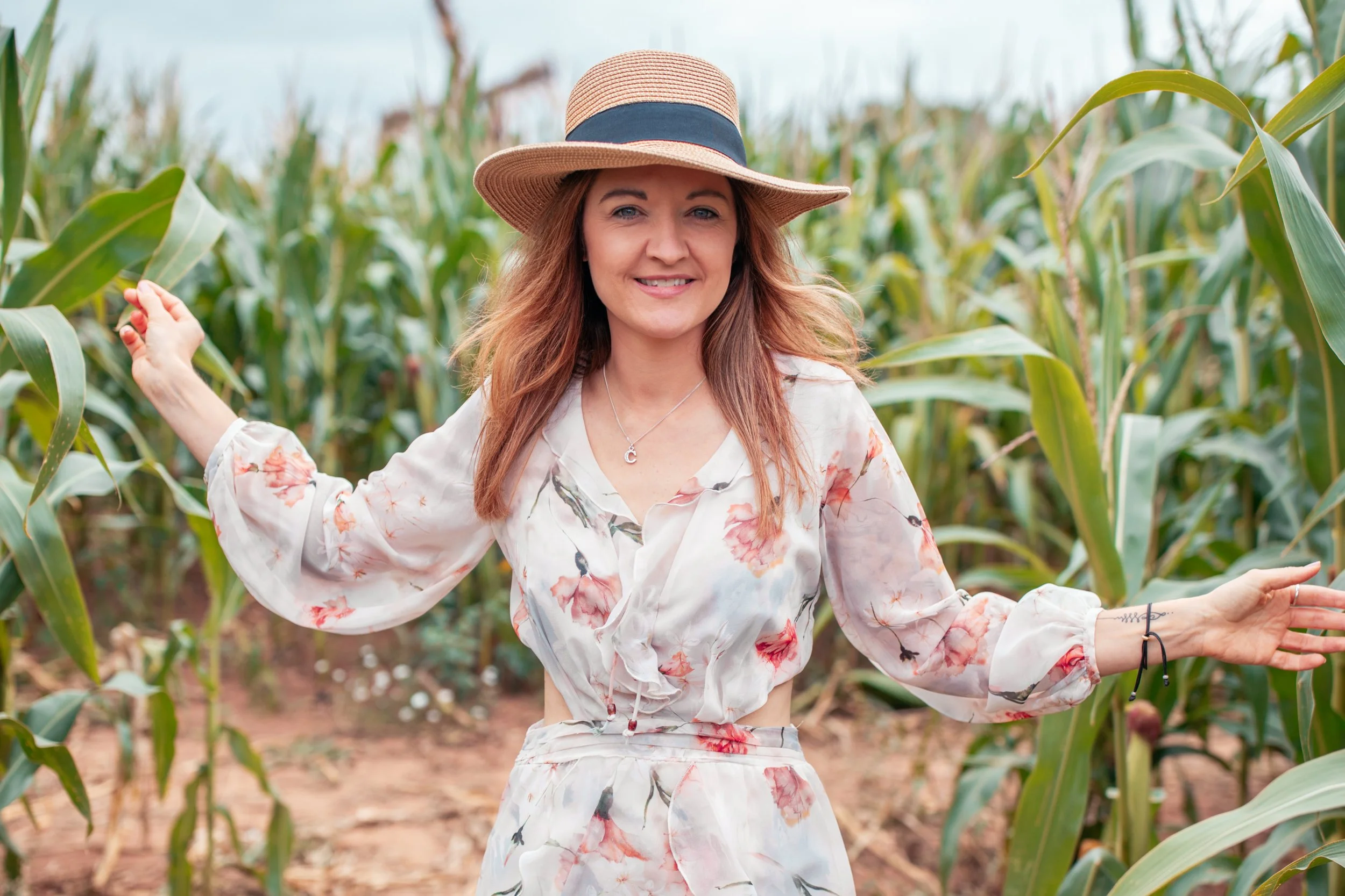 A woman with long wavy hair wearing a wide-brimmed straw hat and a white floral dress standing in a cornfield, smiling with her arms outstretched.  Natural beautiful portrait.
