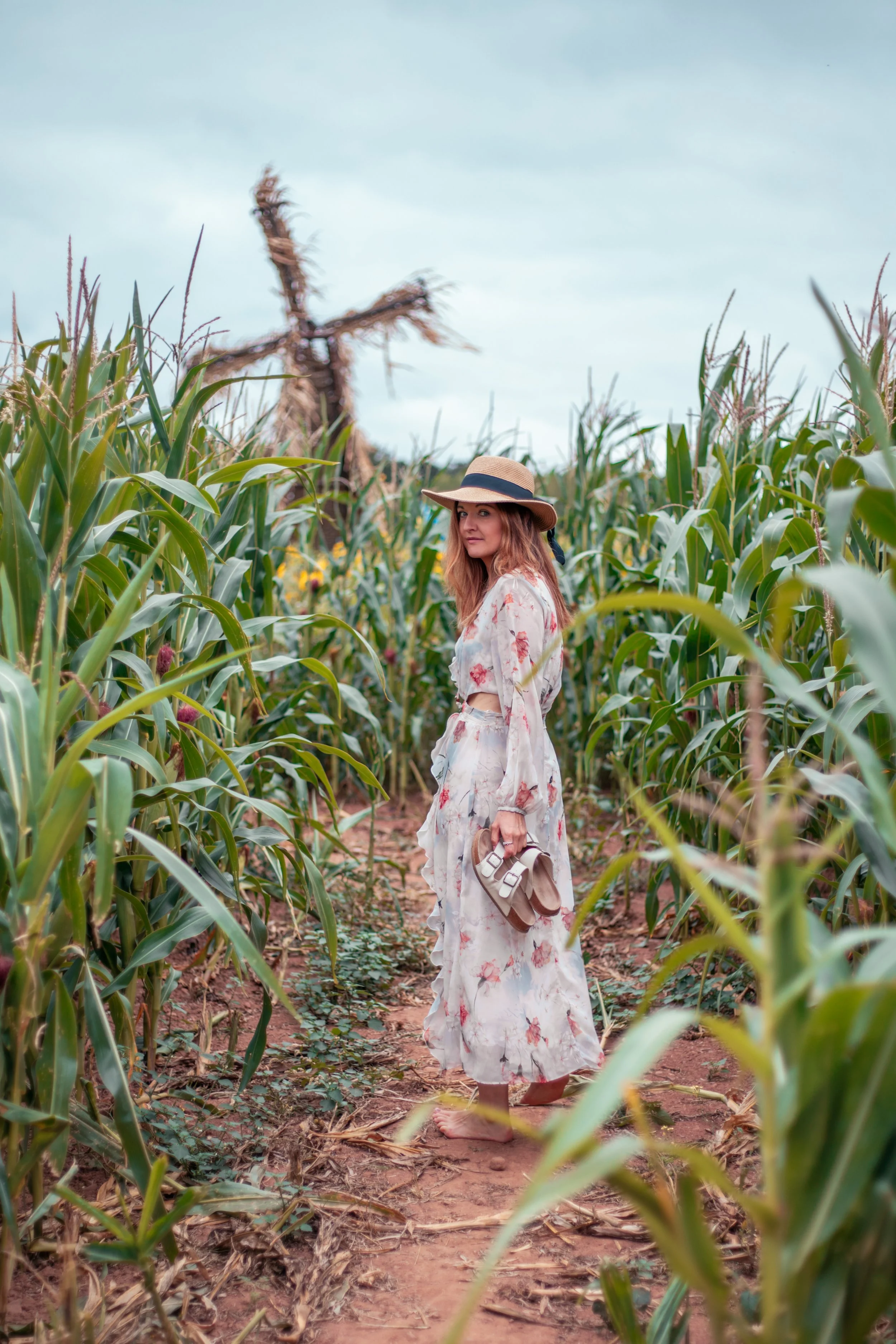 Woman standing barefoot in a cornfield wearing a floral dress and straw hat, holding sandals. Natural beautiful portrait.
