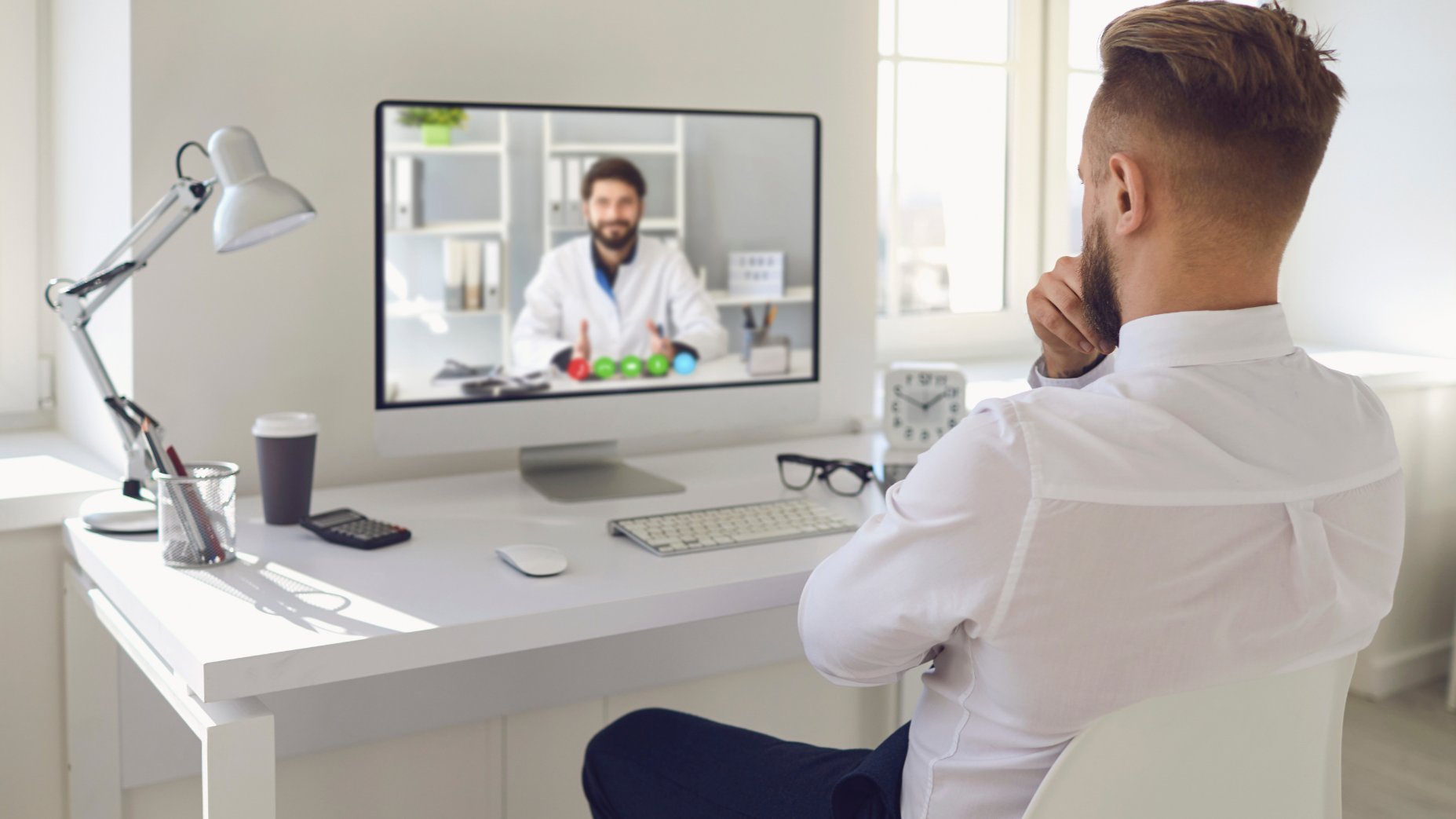 A man in a white shirt sitting at a desk in a bright office, video chatting with a doctor in a white coat on a computer screen.