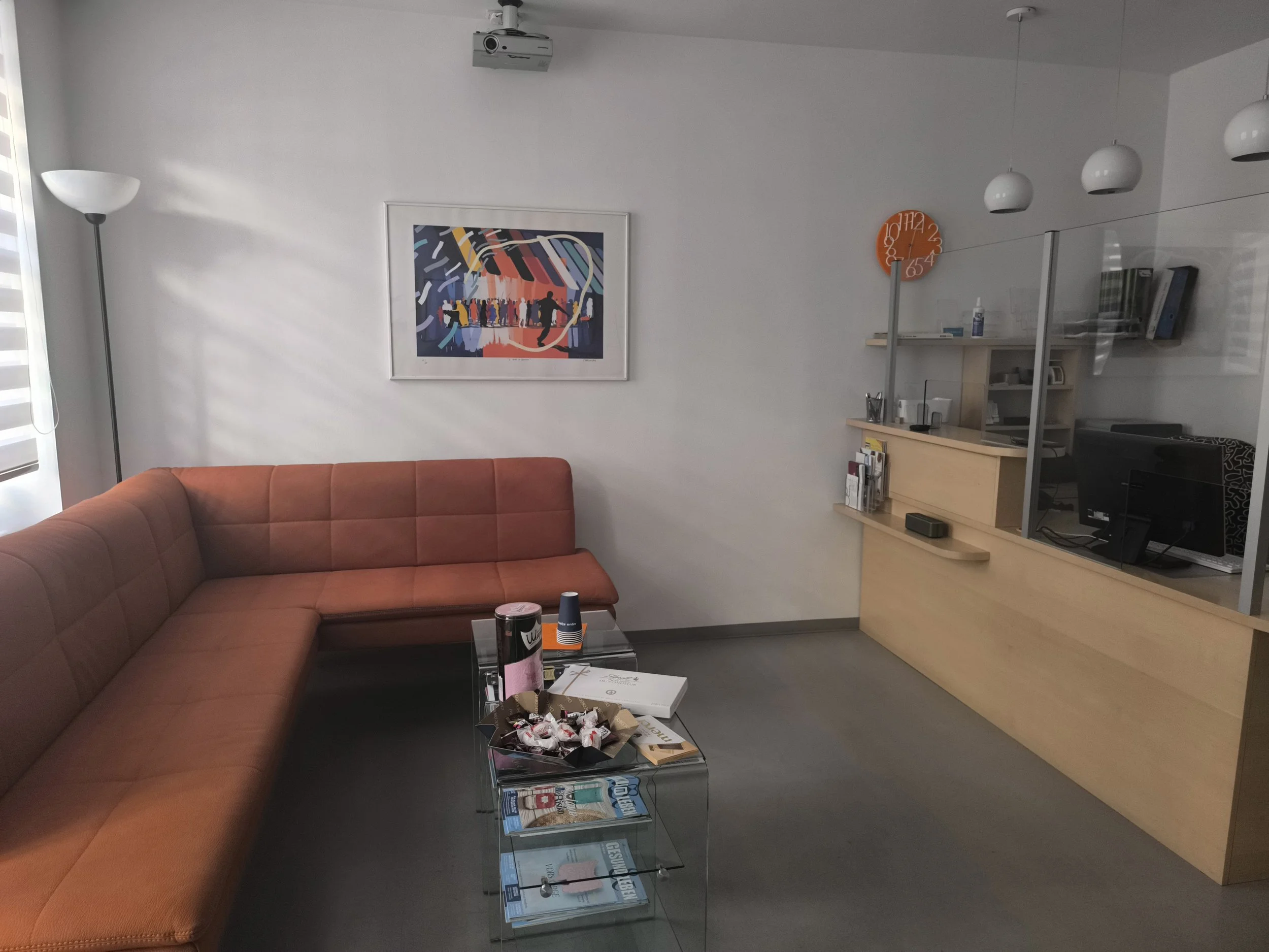 Ein modernes Wartezimmer mit einer großen orangefarbenen Ledercouch, einem Glas-Couchtisch mit Kaffee und Kuchen, einem wandhängenden Kunstwerk, einem orangefarbenen Wandthermometer, einem Empfangstresen mit Computer und Büchern, und einer weißen Stehlampe neben einem Fenster.