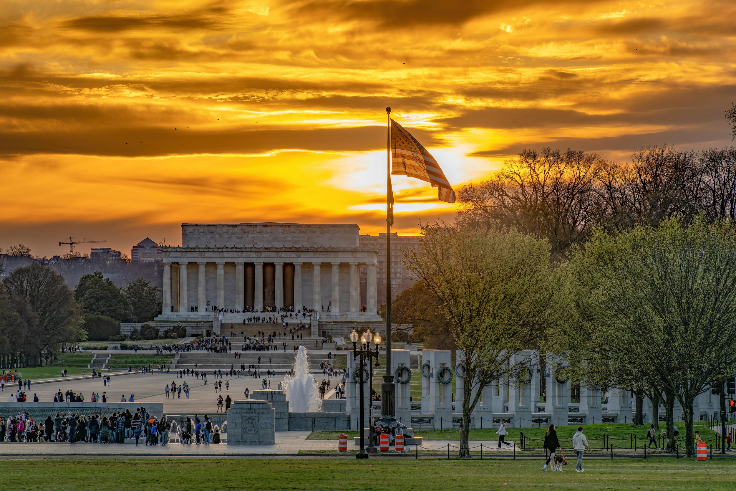 The Lincoln Memorial in Washington, D.C. at sunset with an American flag in the foreground, surrounded by trees and visitors.