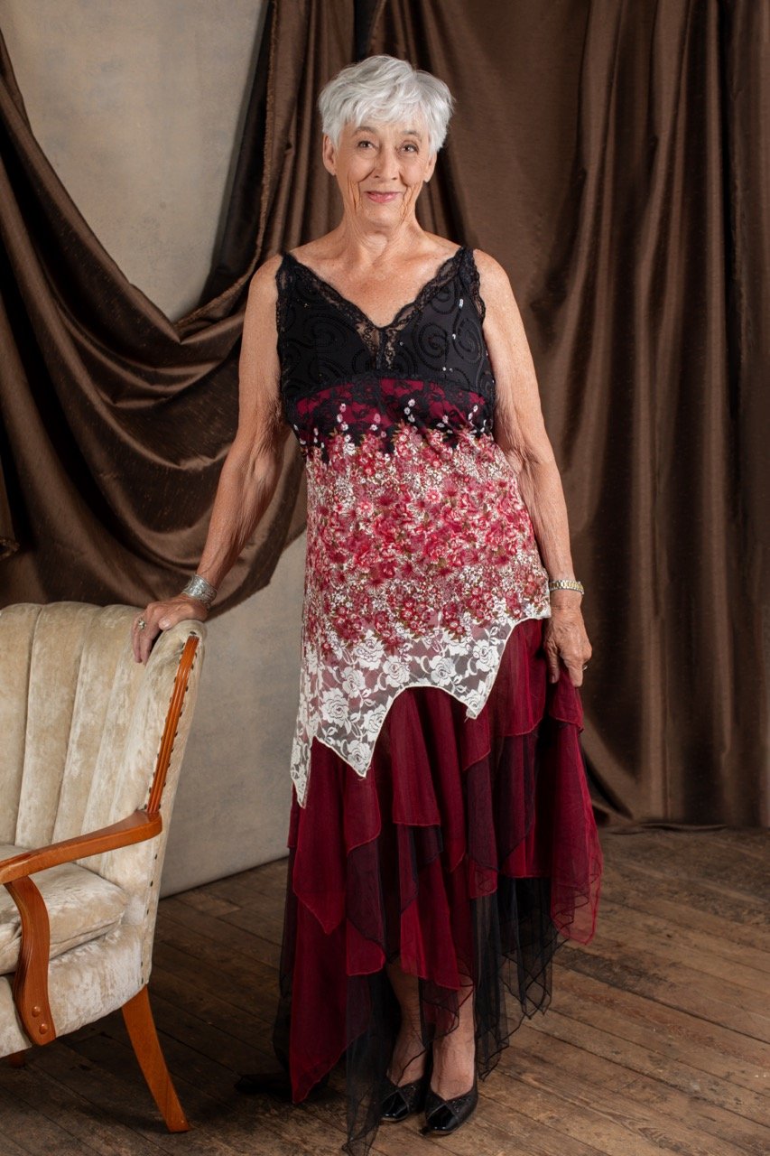 An elderly woman with short white hair stands indoors in front of brown drapes. She is wearing a black, red, and white floral dress with lace and sheer fabric, paired with black heels. She has a silver bracelet on her left wrist and is smiling gently.
