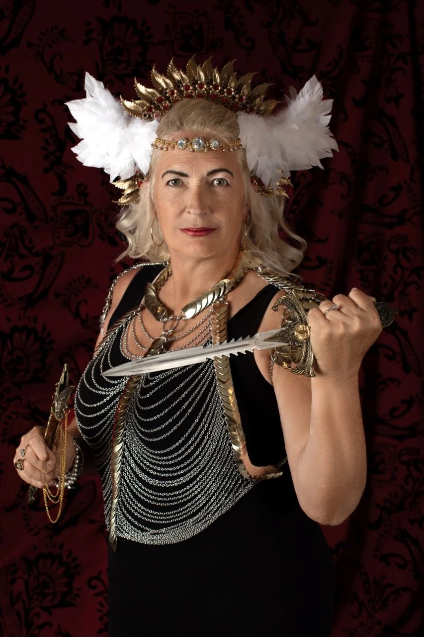 An older woman with long gray hair wearing a regal costume, including a headdress with gold, red, and white feathers, a black dress, and layered necklaces. She holds a sword and wears an expression of confidence against a dark red decorative background.