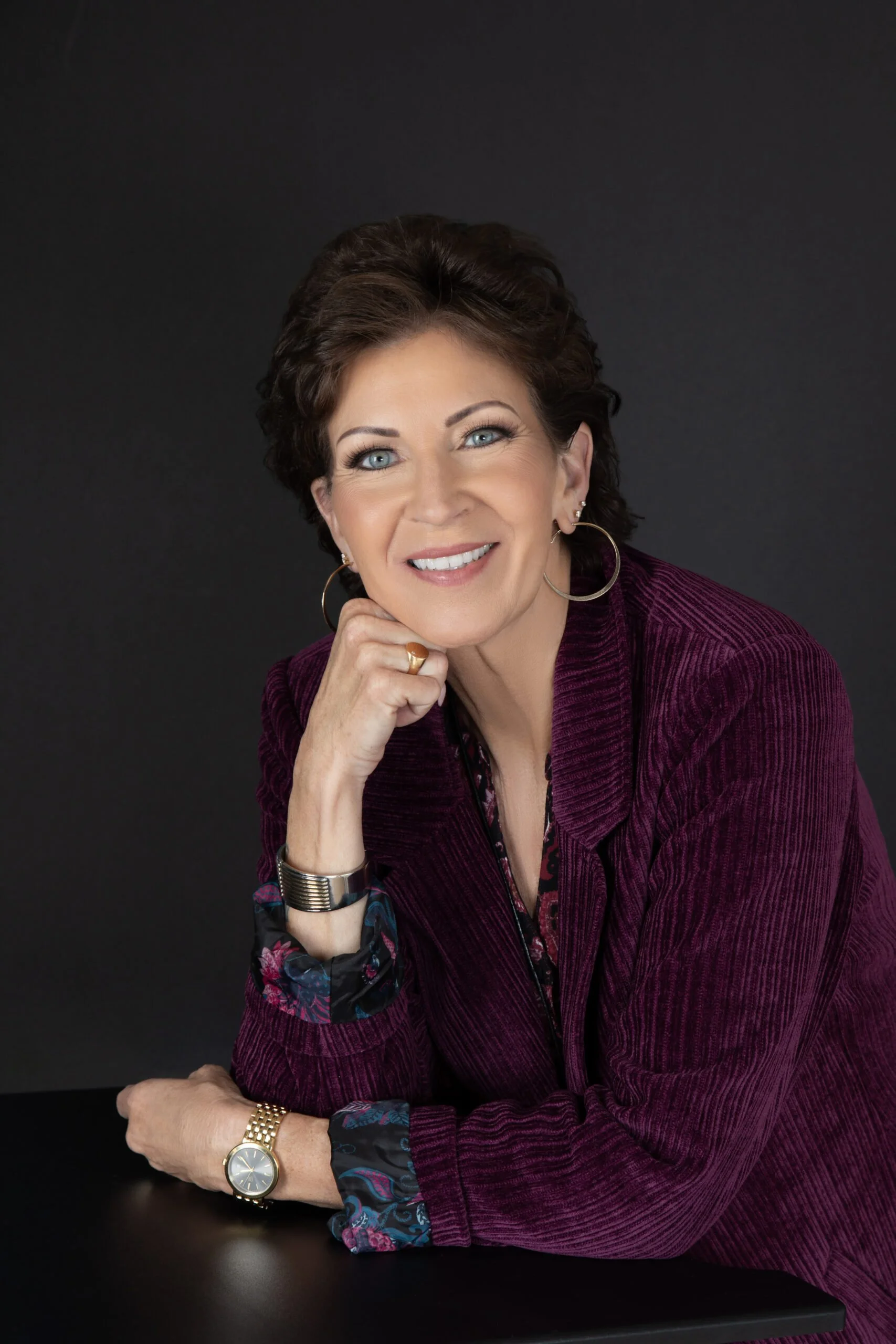 Portrait of a woman with short brown hair and blue eyes, wearing a maroon blazer with floral accents on the cuffs, gold watch, and hoop earrings, smiling and resting her chin on her hand against a dark background.