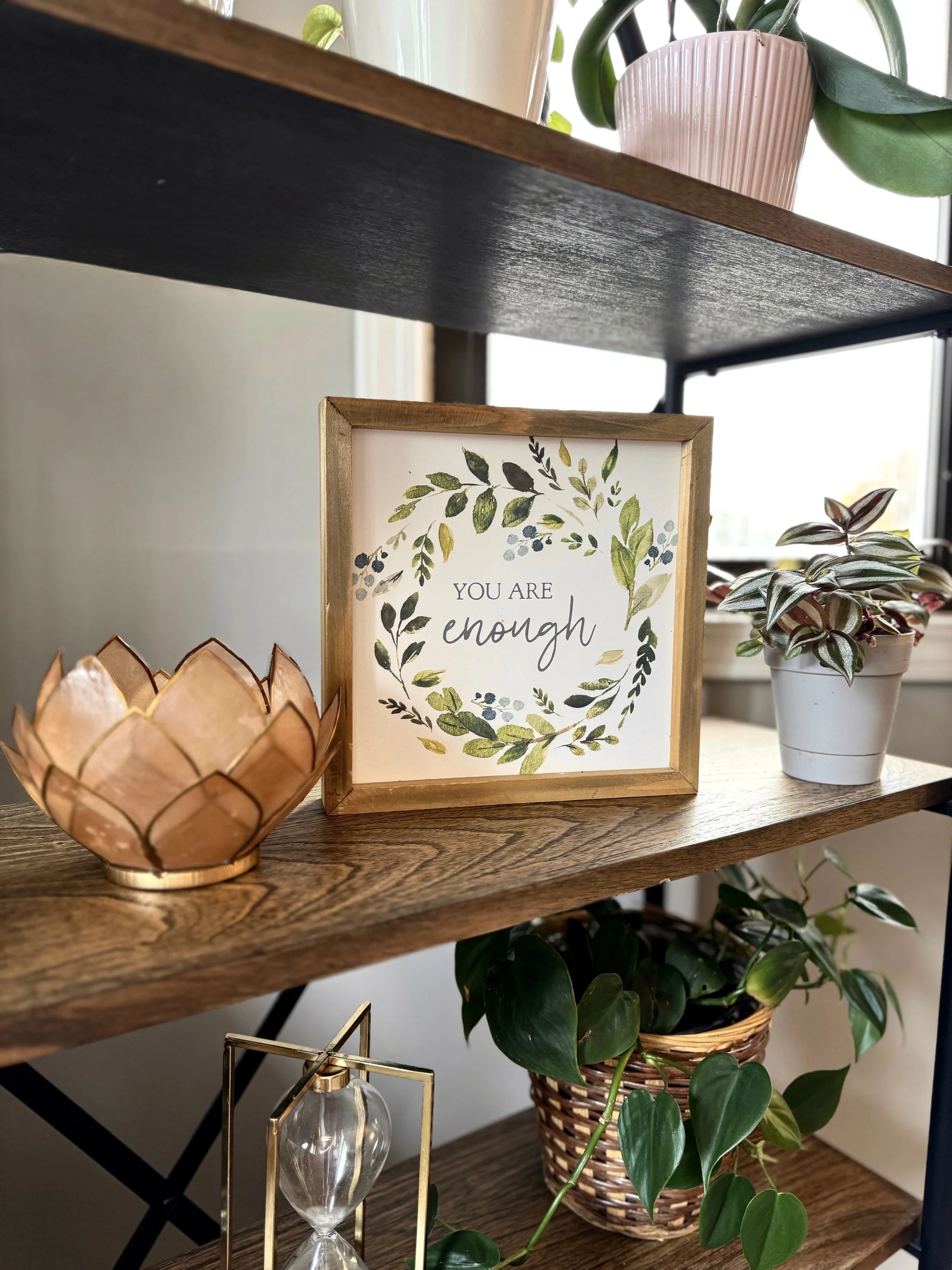 A wooden shelf with a black painted surface holding a pink striped planter with a green houseplant, a framed sign that says 'You are enough' with a leafy wreath design, and a copper lotus-shaped candle holder. Other green plants are visible below and behind the shelf.