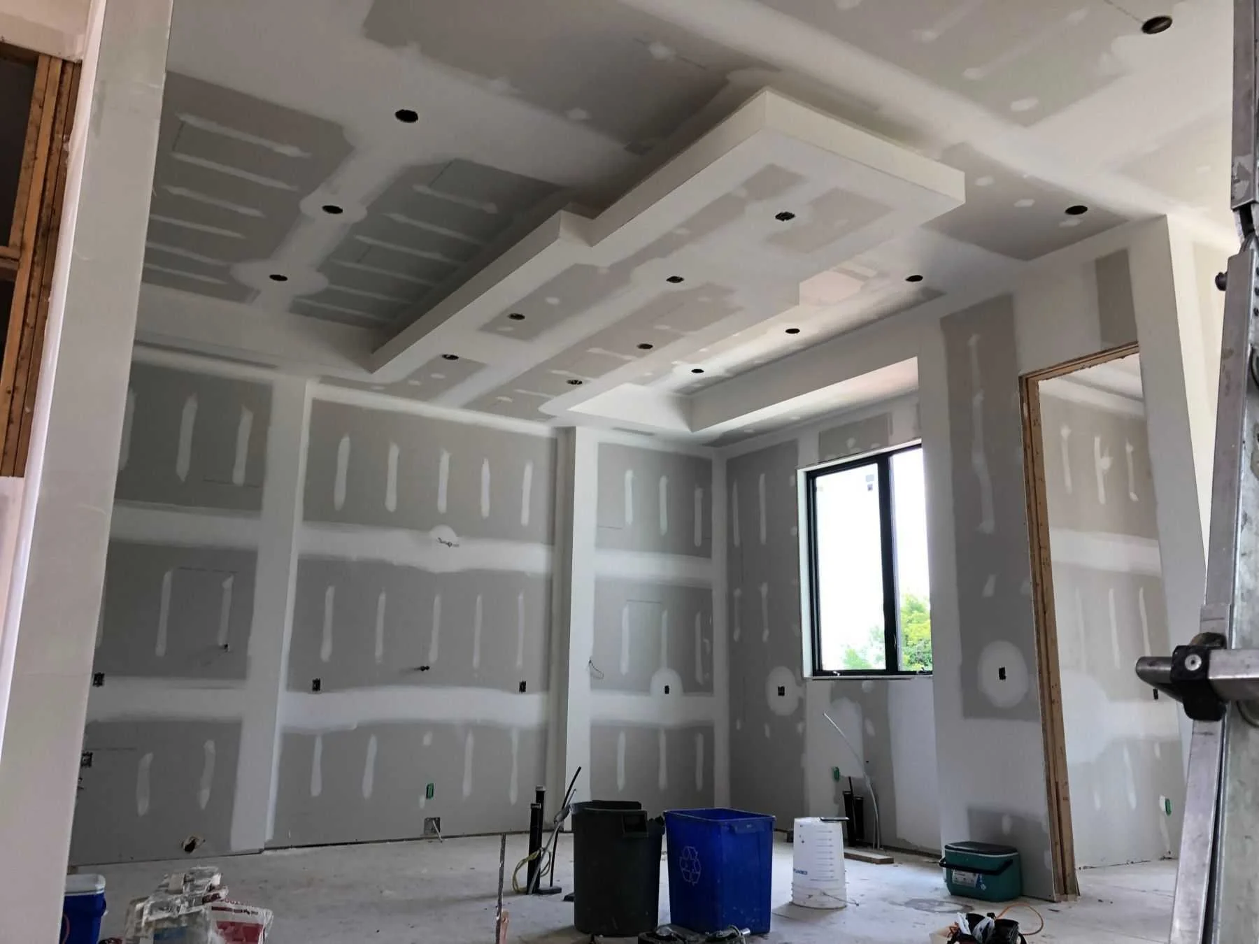 Interior of a room under construction with drywall walls and ceiling, a window, and construction tools and supplies on the floor.