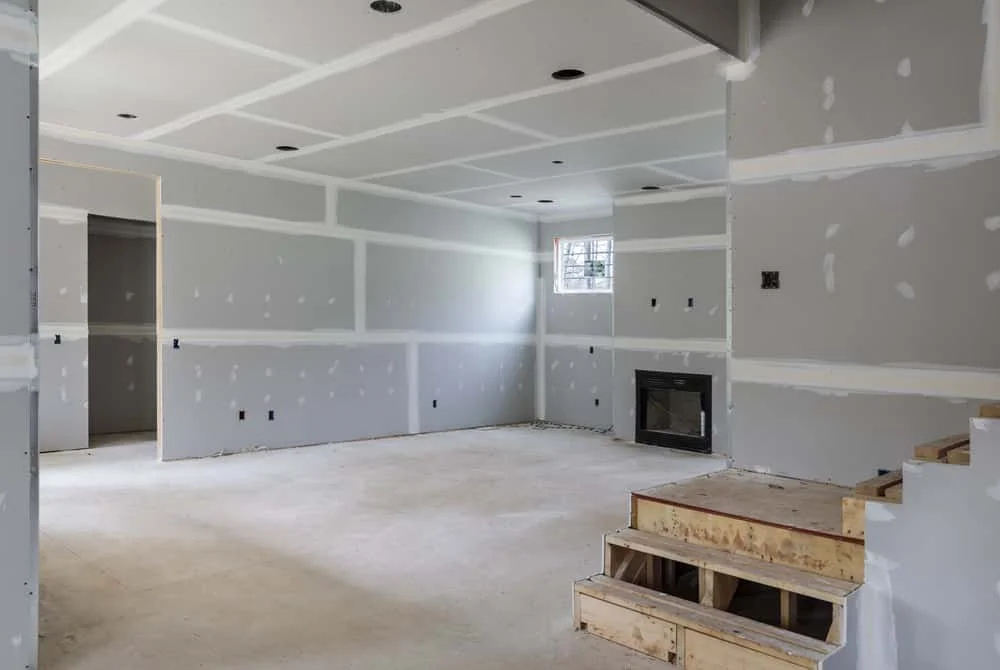 Interior of a room under construction with drywall partially finished, a fireplace, a window, and unfinished wooden stairs.