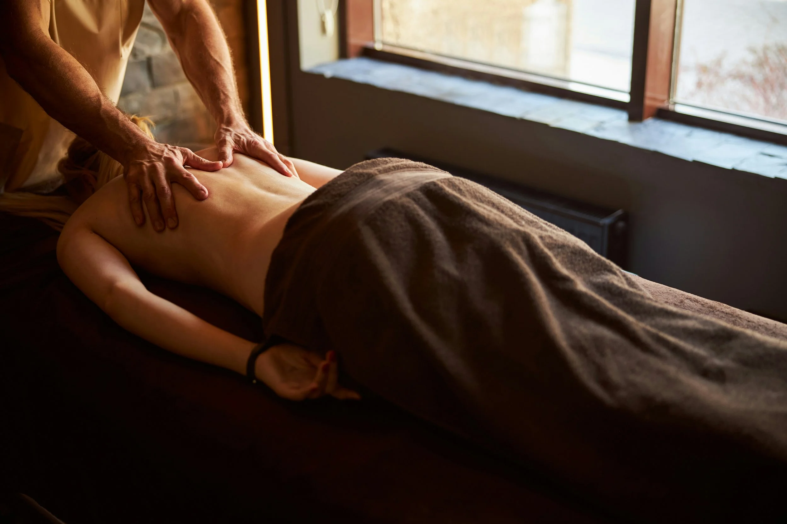 A person receiving a back massage from a massage therapist in a spa or massage room with large windows letting in natural light.