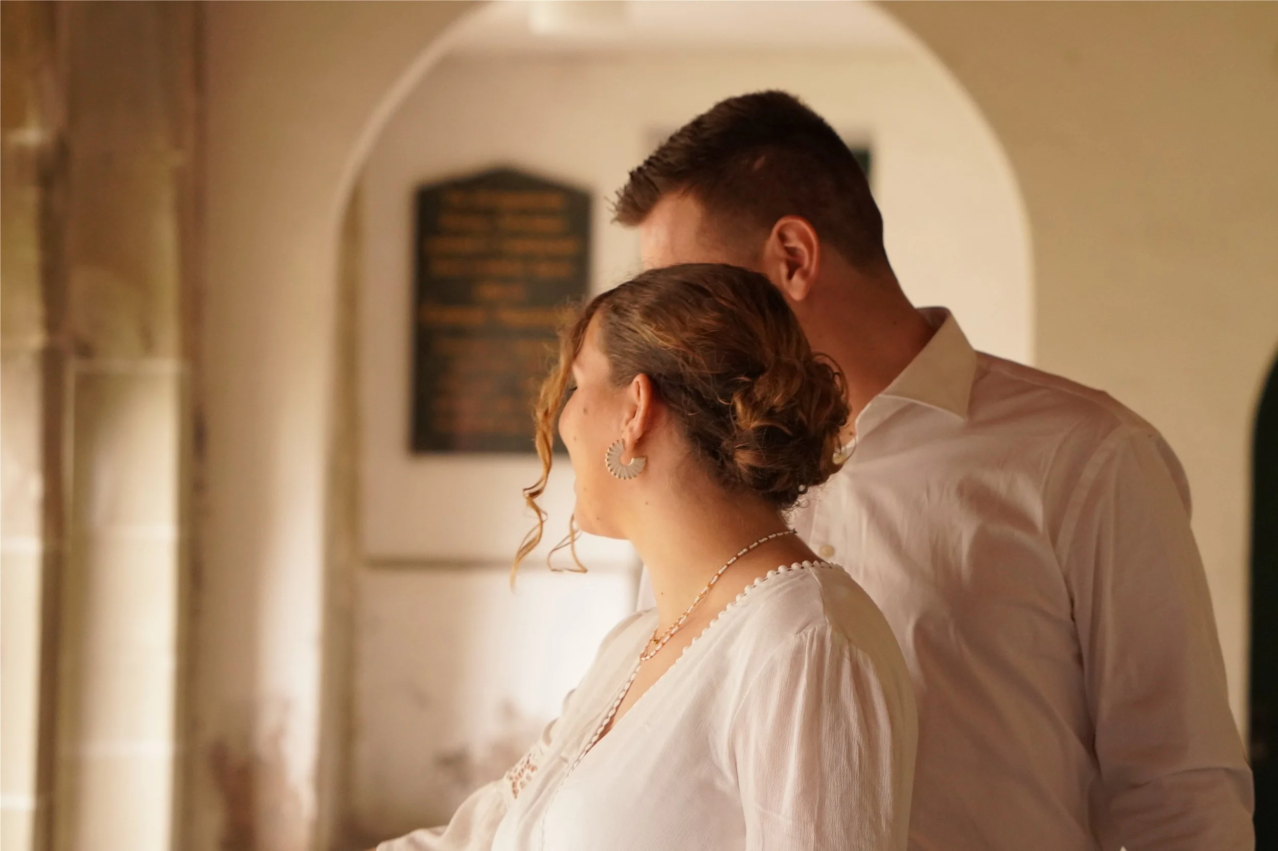 Ein Mann und eine Frau stehen eng beieinander in einem Innenraum mit warmem Licht, ihre Gesichter sind nicht sichtbar, nur die Seite ihres Gesichts und ihre Profile sind zu sehen.