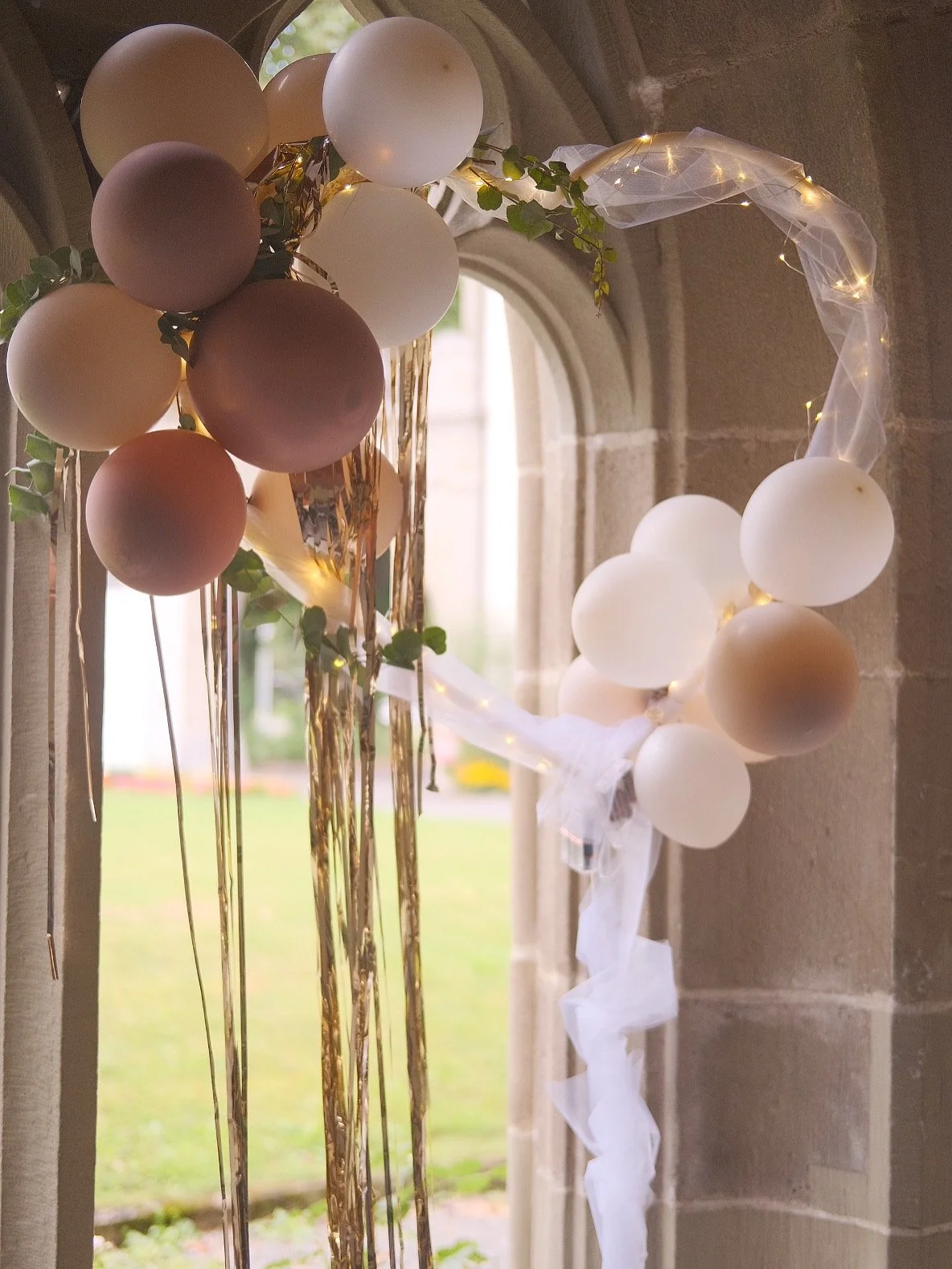 Ein Dekorationsbogen mit weißen, rosa und bronzefarbenen Ballons, drapierter weißer Tüllstoff mit Lichterketten, grünen Blättern und goldenen Streifen, vor einem Fenster in einem steinernen Raum.