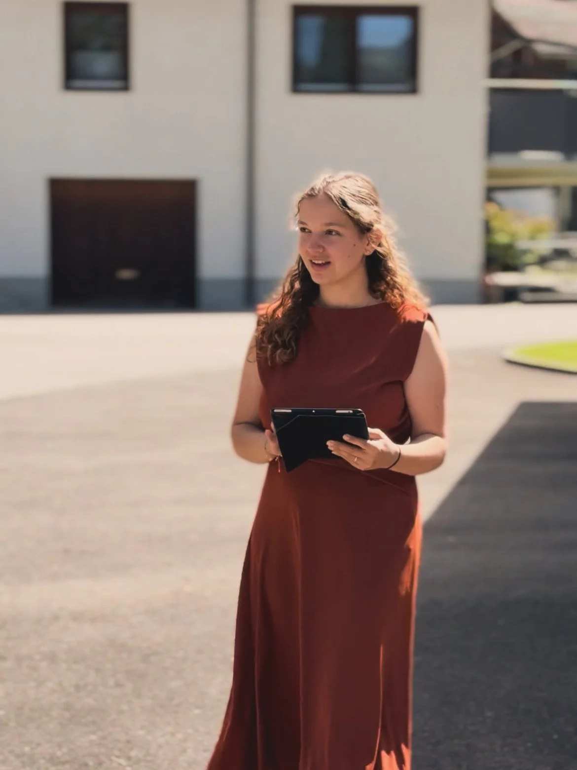 Junge Frau trägt rotes Kleid und hält Tablet im Freien vor modernem Gebäude.