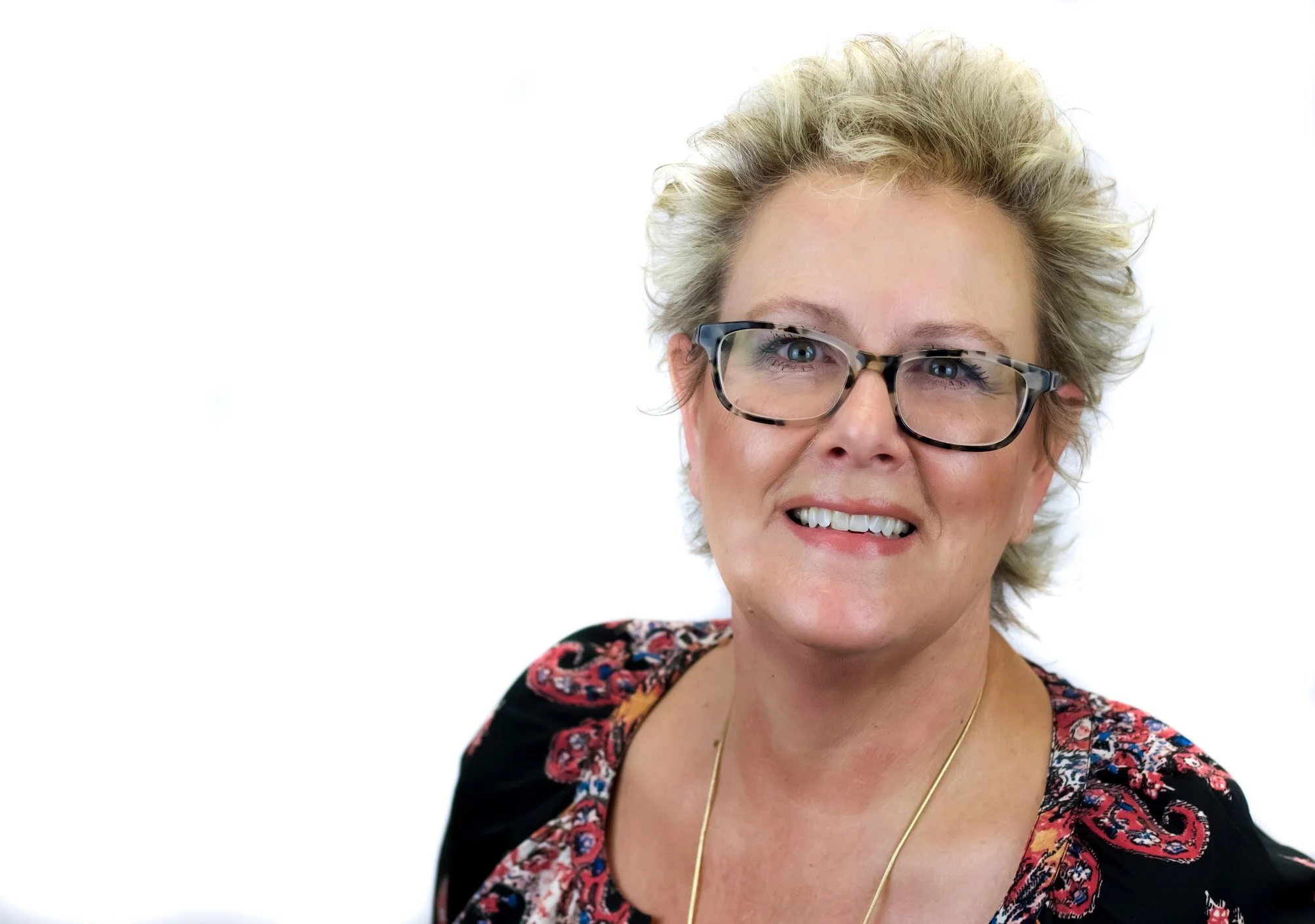 A smiling middle-aged woman with short, curly blonde hair and glasses, wearing a black top with red, gray, and orange patterns and a gold necklace, against a plain white background.