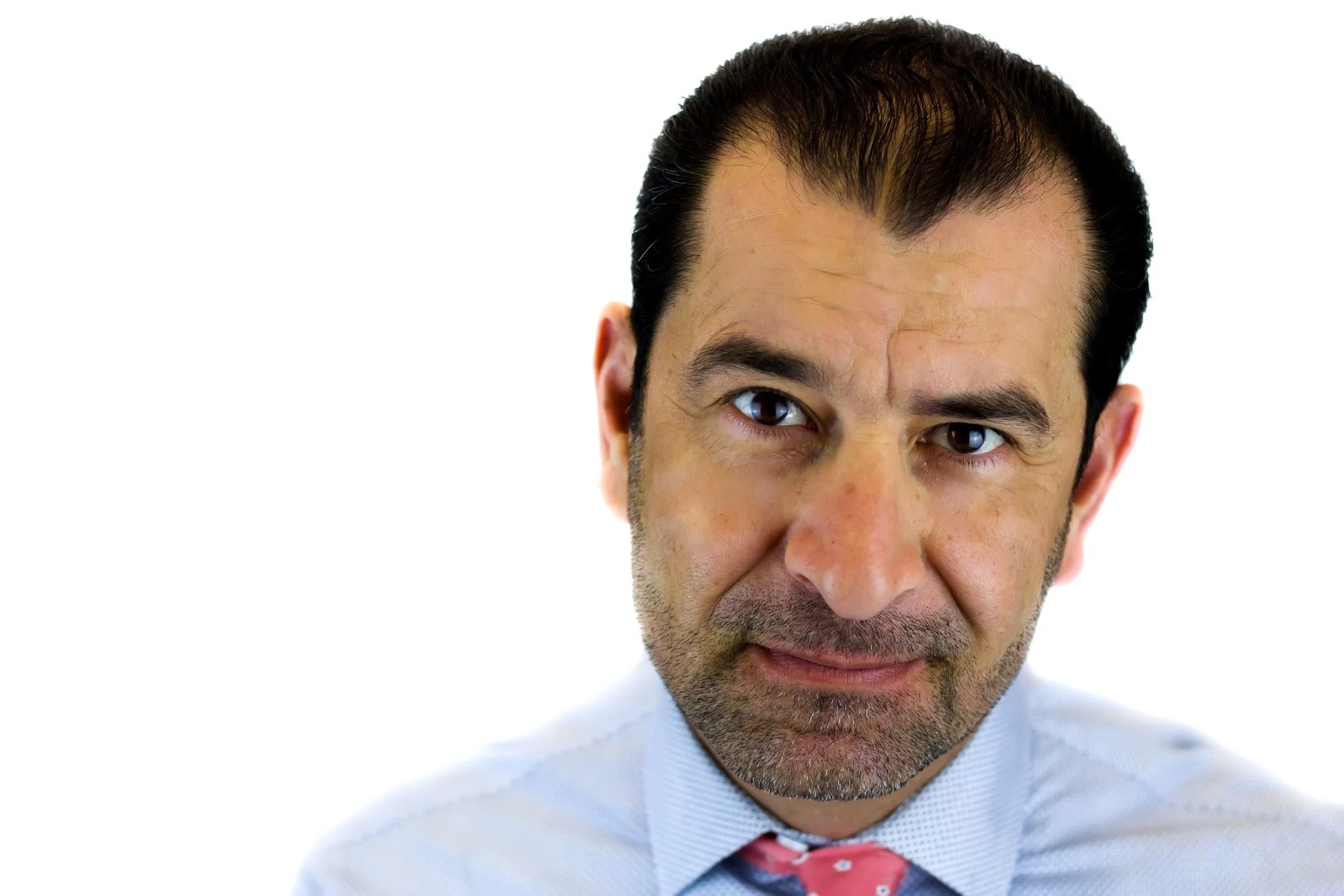 Close-up of a man with dark hair, wearing a collared shirt and a pink tie, looking directly at the camera against a plain white background.