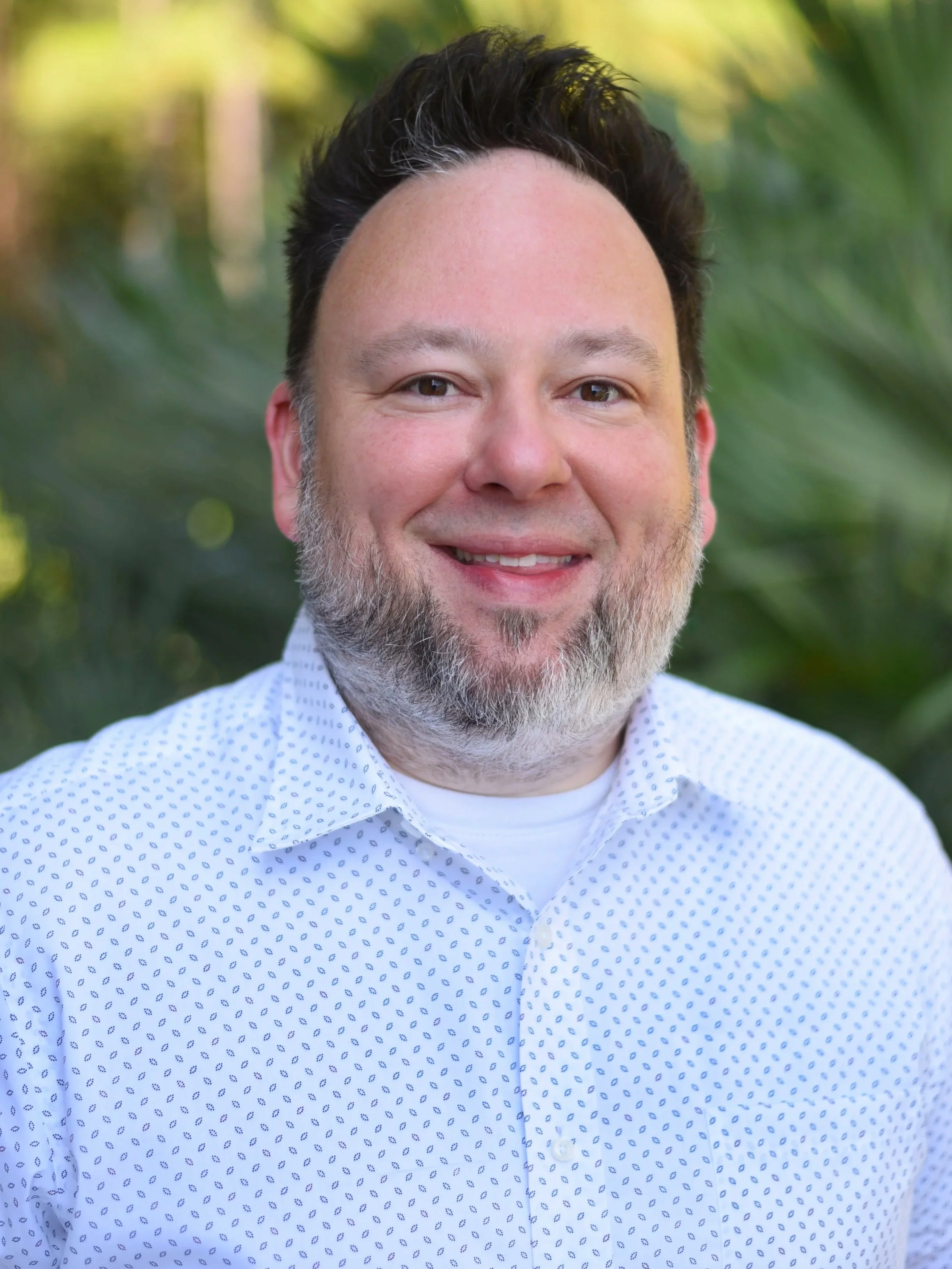 A smiling man with dark hair and a beard, wearing a white patterned shirt, outdoors with green foliage in the background.