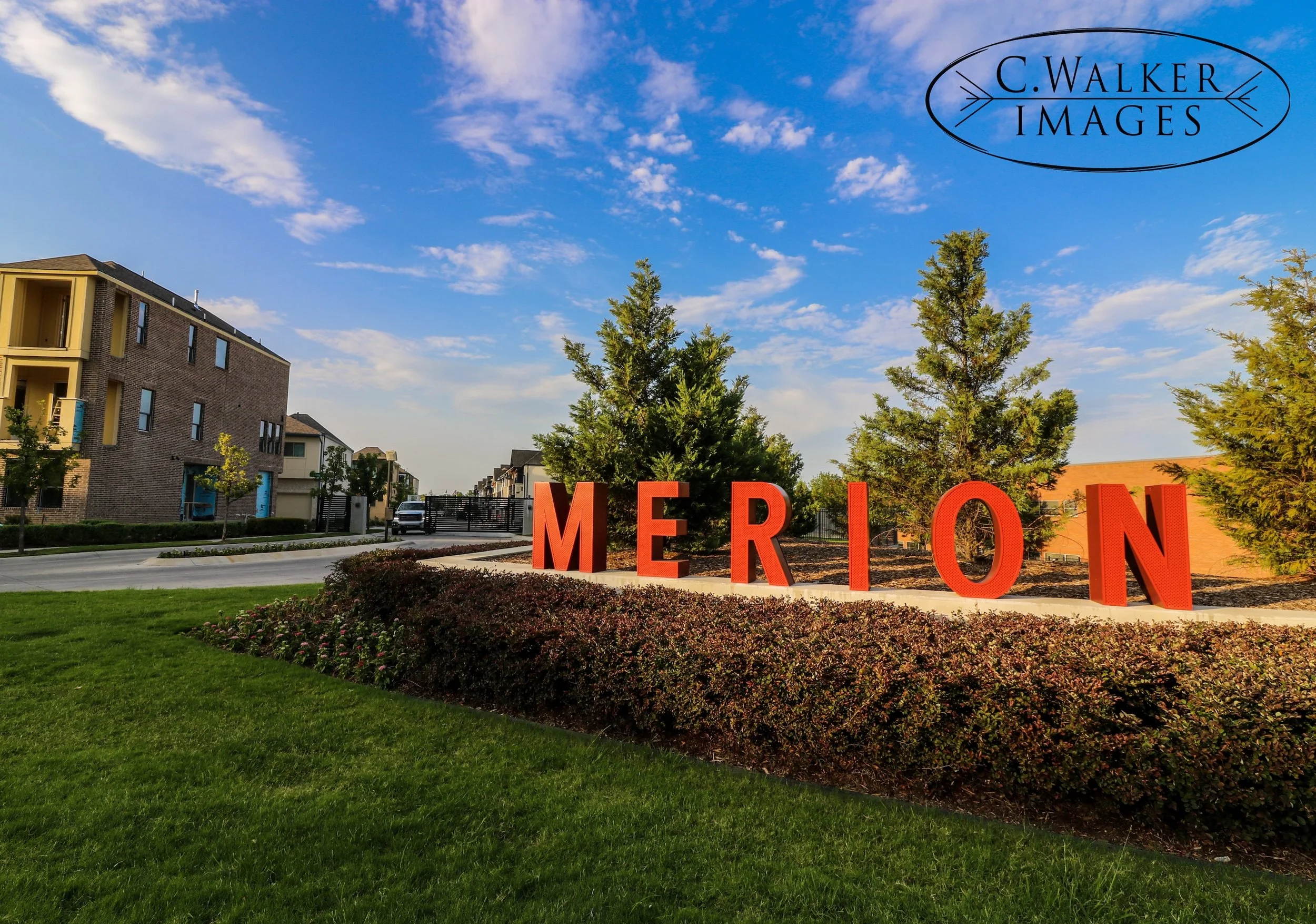 A modern residential area with multi-story buildings, green lawns, trees, and large red letters spelling "MERION" in the foreground under a bright blue sky with some clouds.