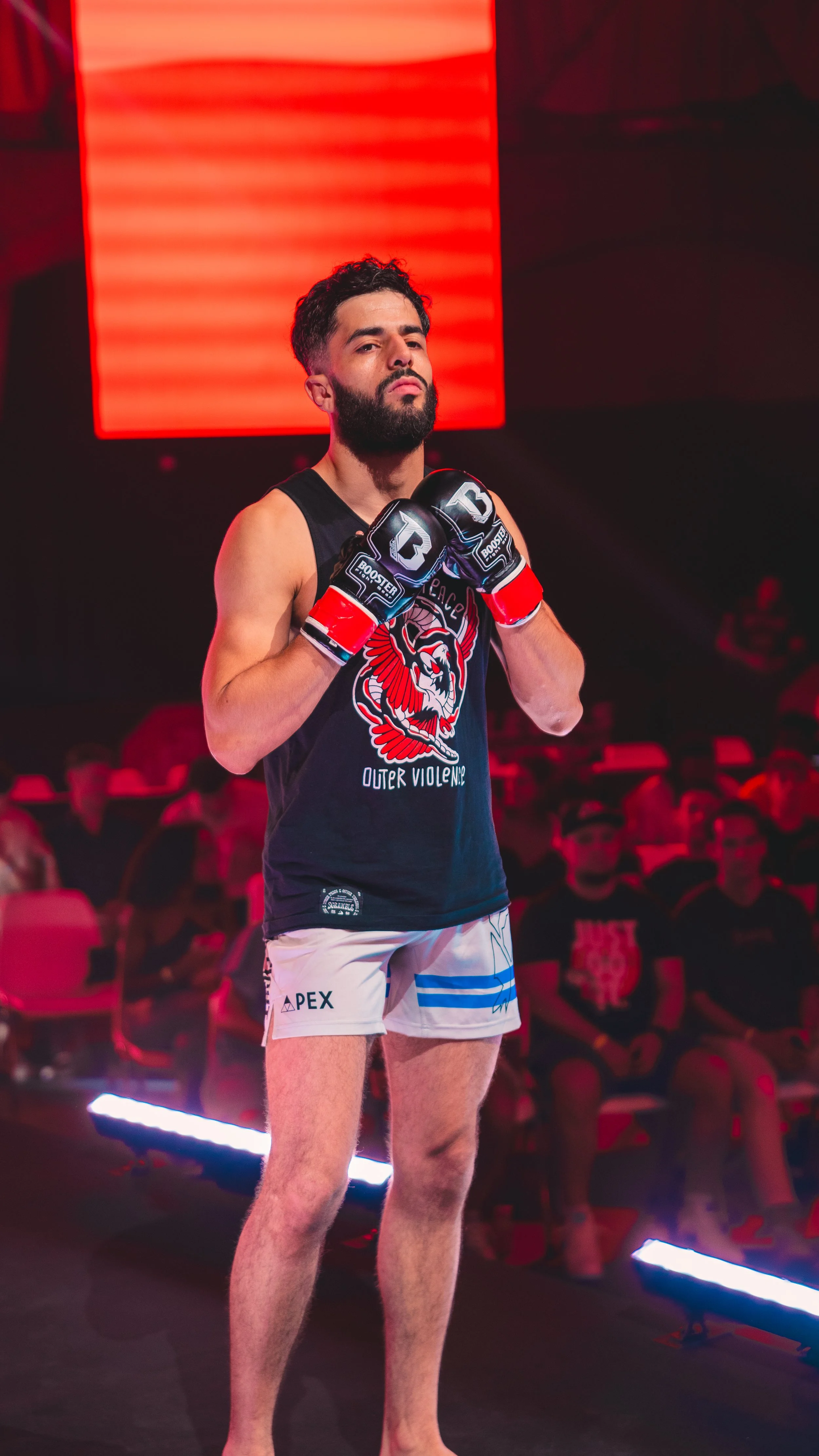 A male MMA fighter stands in the fighting stance at a fighting event, wearing gloves, a black sleeveless shirt, and shorts.
