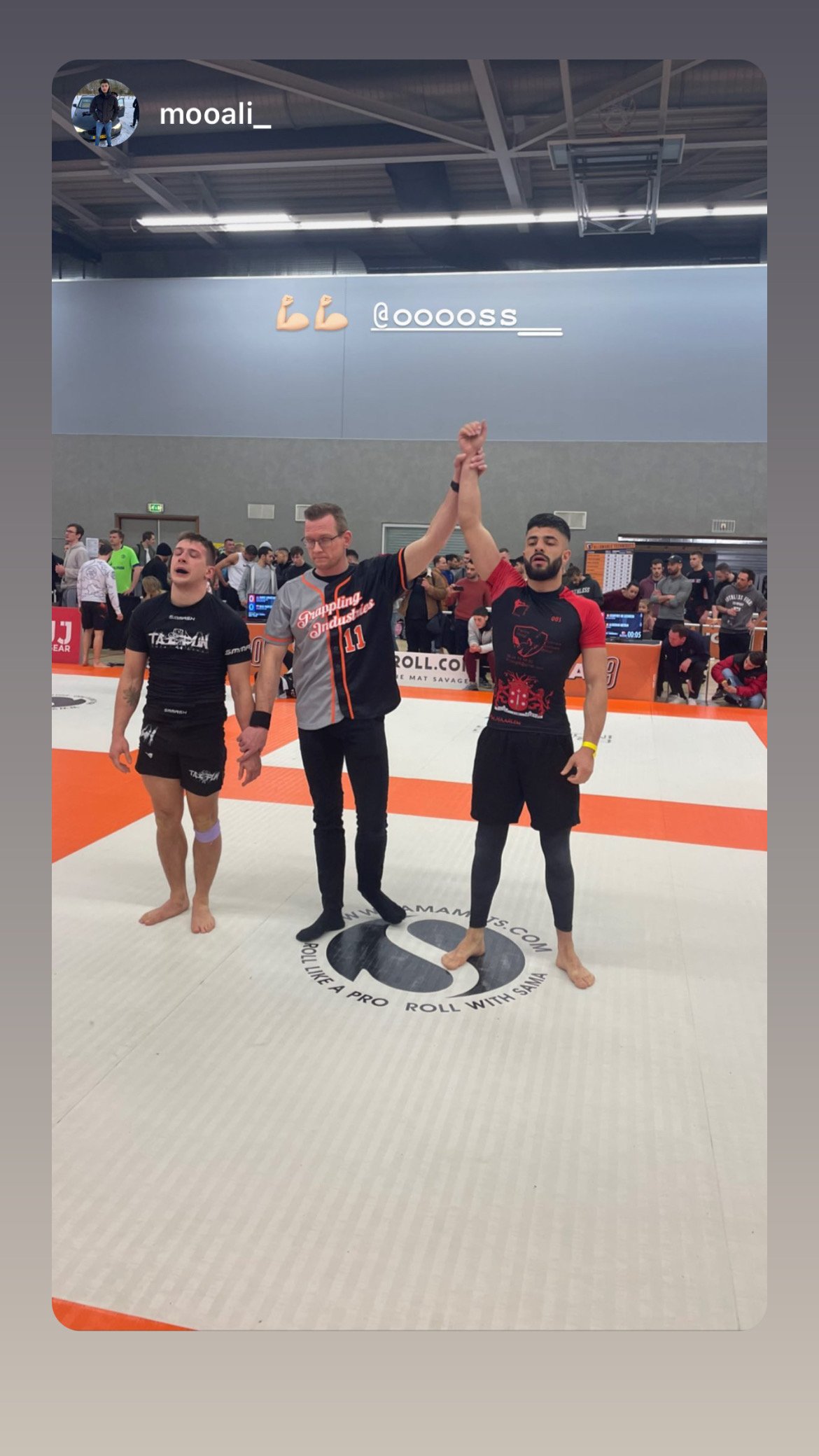 Two male athletes standing on a grappling mat, with a referee in the middle raising the winner's arm, at a martial arts competition. The crowd is visible in the background at an indoor arena.