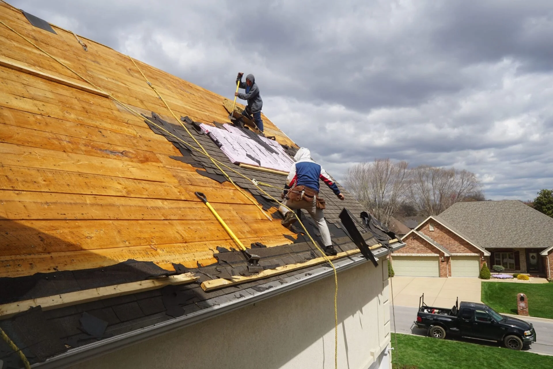 Two workers are repairing a roof on a house, with one on the wooden part of the roof and the other on the shingled section, under cloudy skies.