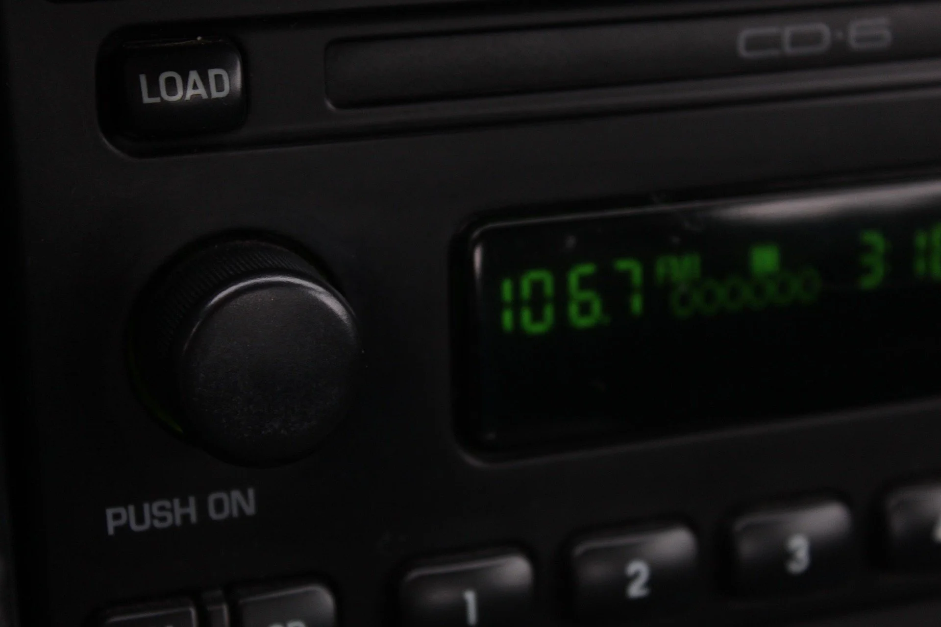 A close-up of a car stereo showing a green digital frequency display with the numbers 106.7 FM. Part of a "LOAD" button and some control knobs are visible.