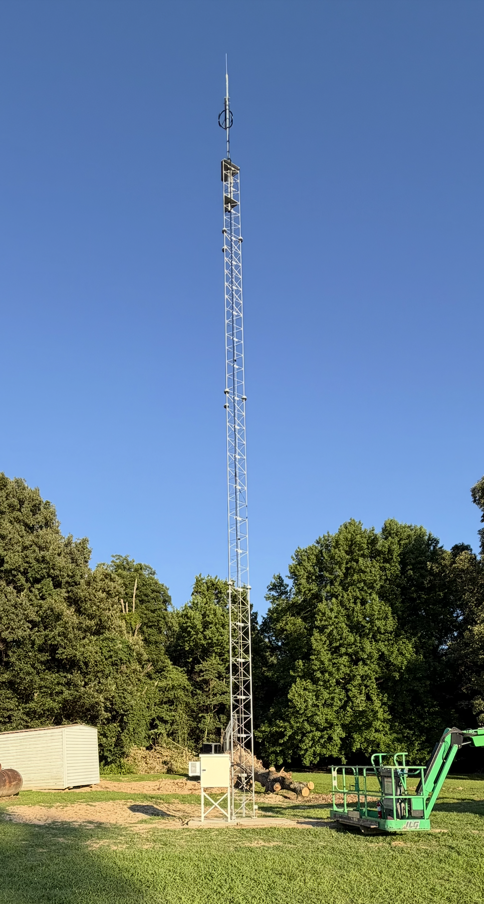 A tall metal communication tower on a grassy area with a clear blue sky overhead and trees in the background.