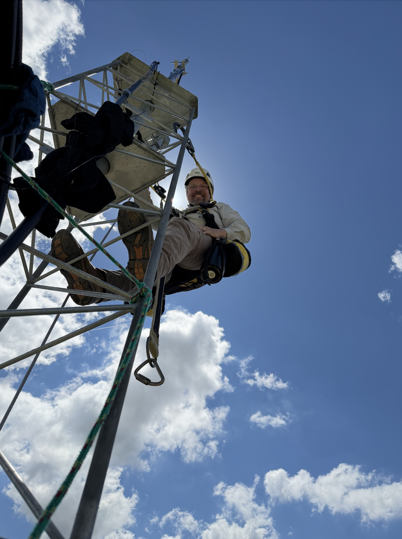 A man wearing a helmet and harness climbing a metal tower or antenna against a blue sky with clouds, with safety ropes and equipment visible.