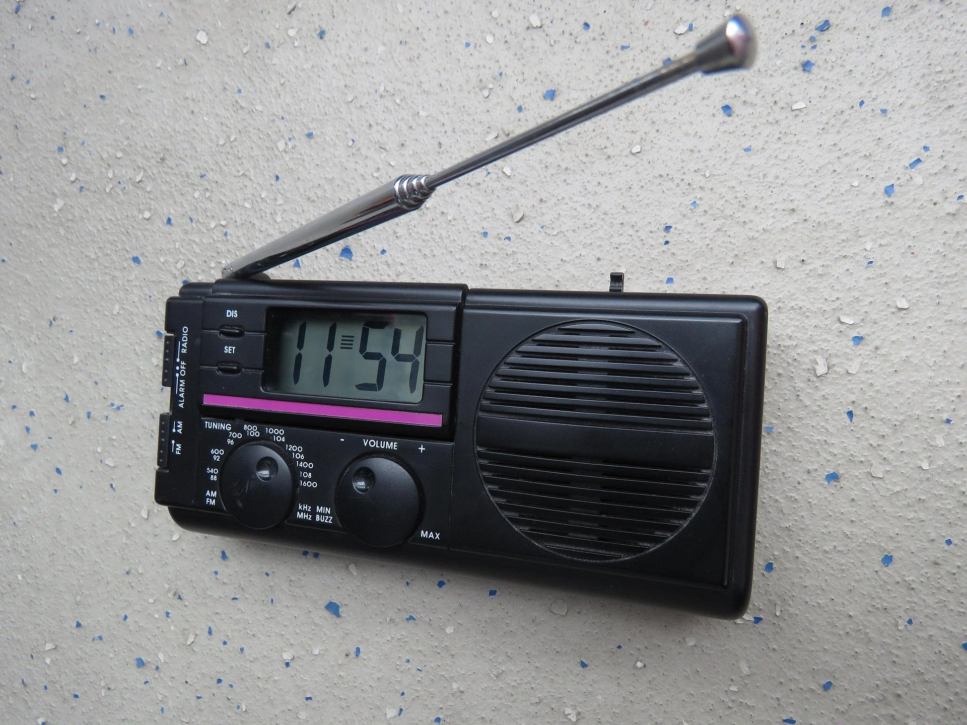 A black portable radio with an extendable antenna displaying the time 11:54, placed against a speckled white and blue textured wall.