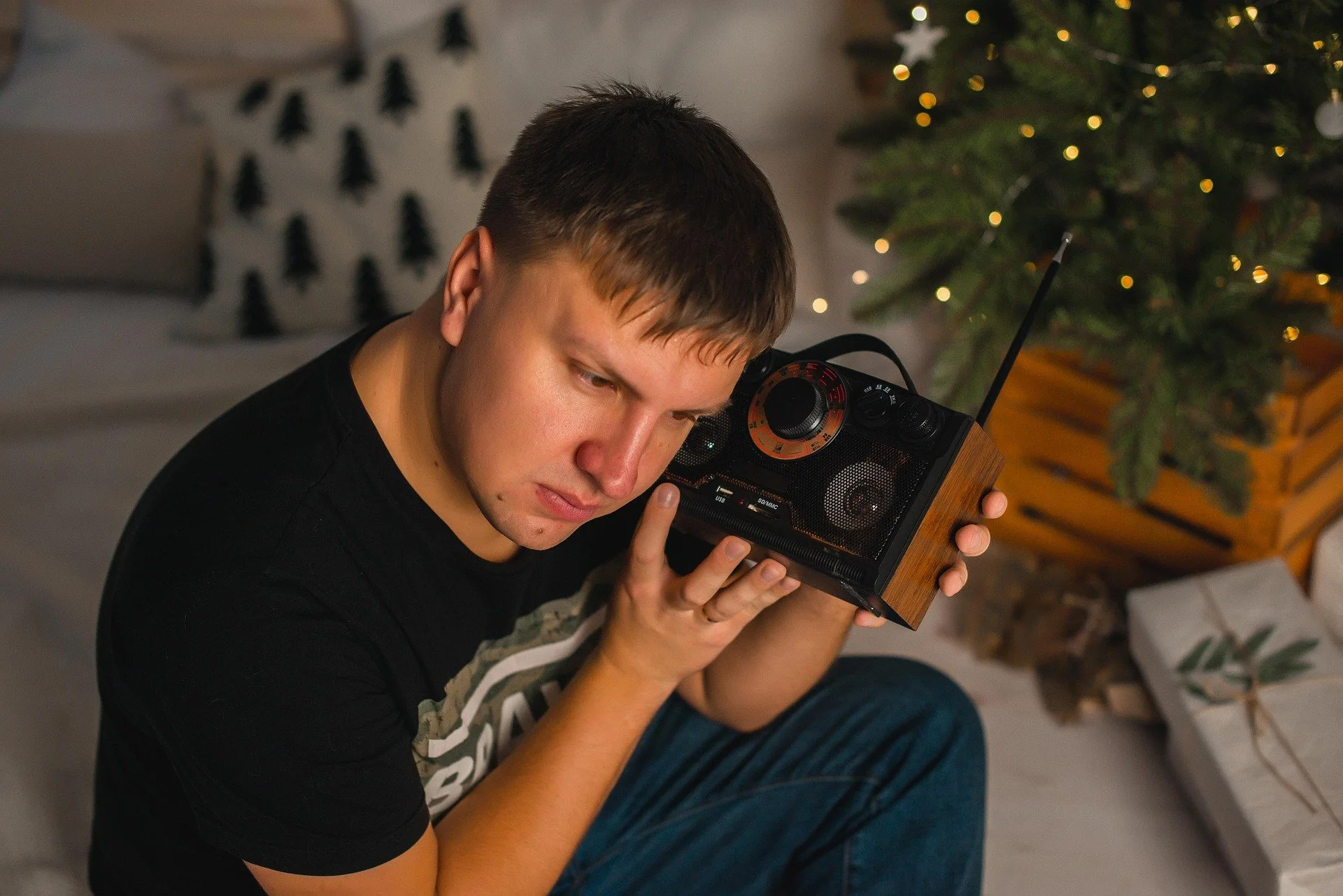 A young man with short brown hair holding a portable radio close to his face, sitting in a room with a decorated Christmas tree and white pillows with black Christmas tree patterns in the background.