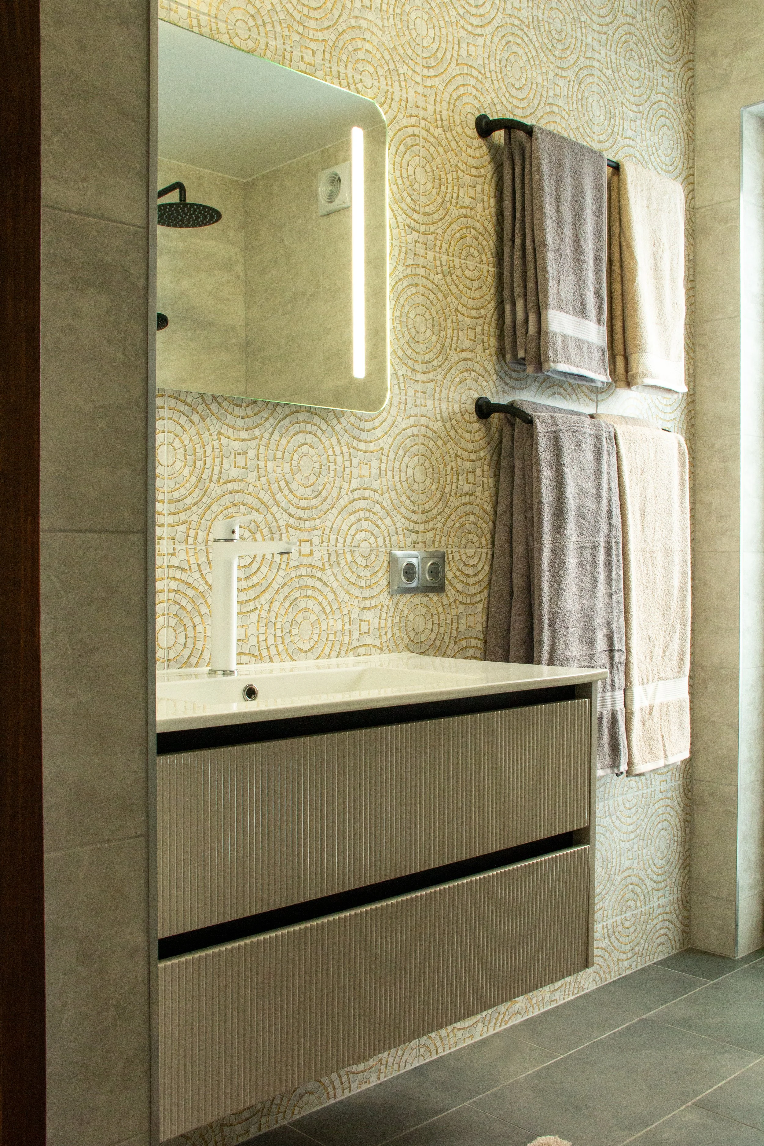 Bathroom with beige patterned wallpaper, a rectangular mirror with an LED light strip, a white sink with a modern white faucet, two gray towels and one beige towel hanging on black towel rods, a window on the right, a wall outlet, and a partially vis