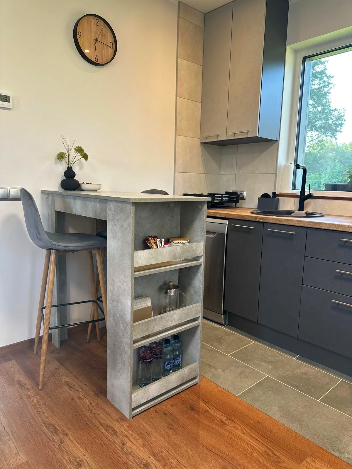A modern kitchen with a wall clock, a small counter with a vase of flowers, dark gray lower cabinets, beige upper cabinets, a window, a sink, and some water bottles underneath a shelf.