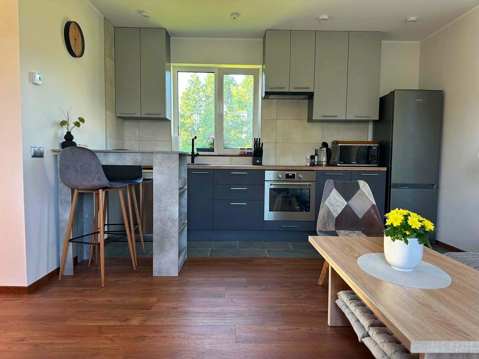 Kitchen with gray upper cabinets, black lower cabinets, a window above the sink with trees outside, stainless steel oven, microwave, refrigerator, and bar area with two modern chairs; wooden dining table with a vase of yellow flowers.
