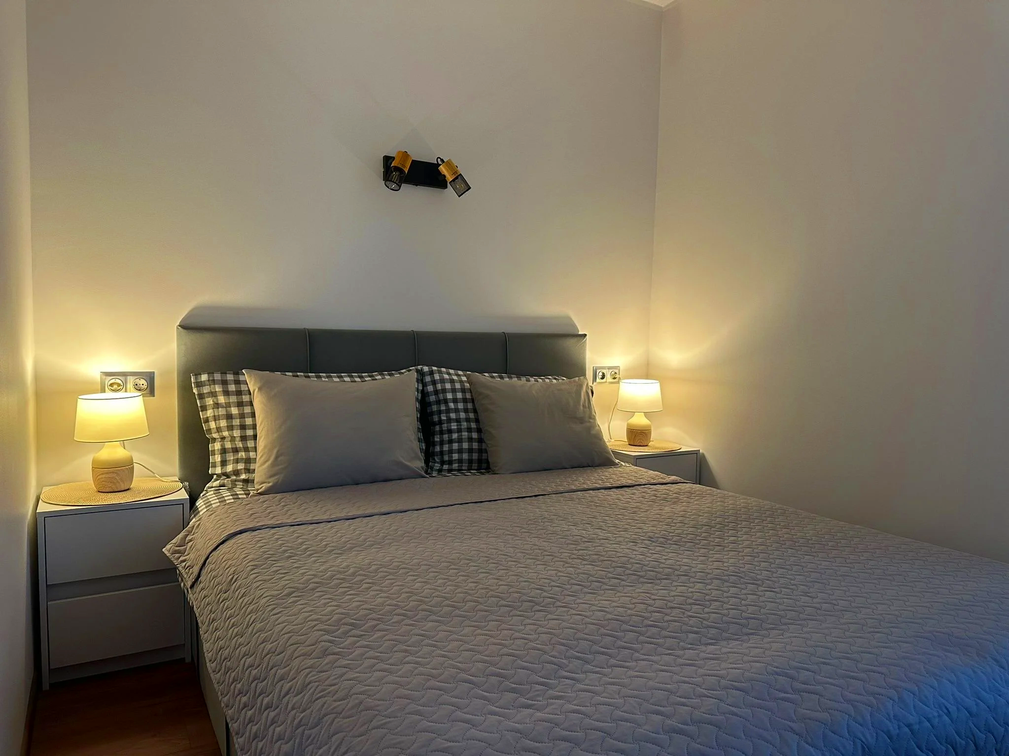 A neatly made bed with a gray headboard, beige quilt, and four pillows, two in beige pillowcases and two with black and white checkered pillowcases. Two bedside tables with beige lamps are on each side of the bed. The wall above the bed is empty exce