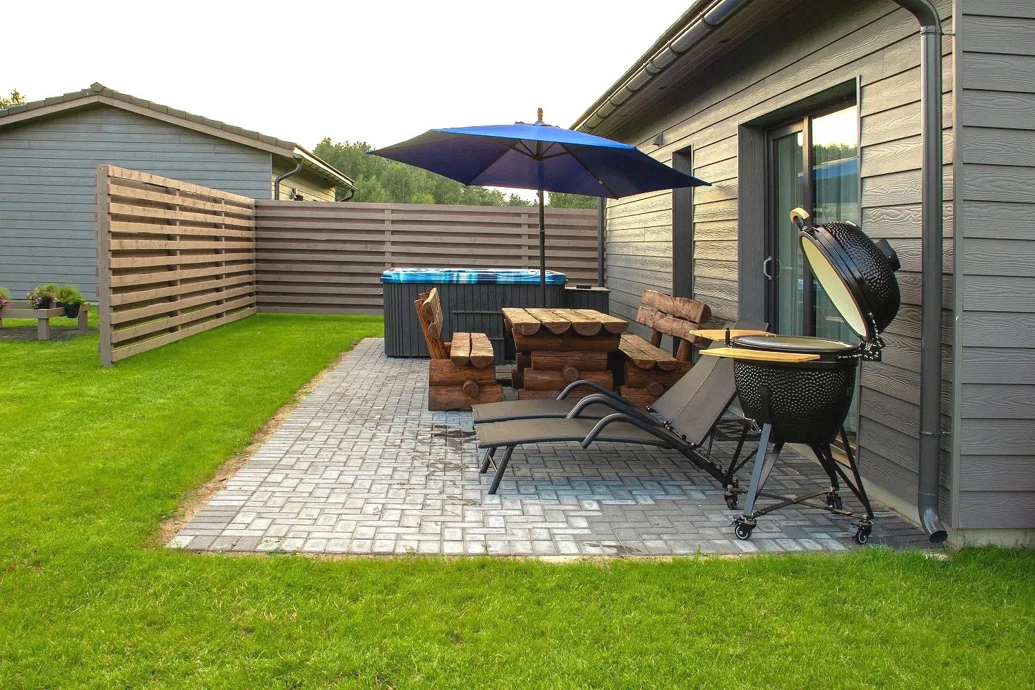 Backyard patio with lounge chairs, wooden table and benches, parasol, hot tub, grill, and a lawn, surrounded by wooden fencing and gray houses.
