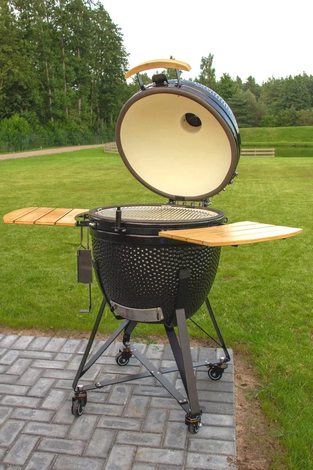 A black portable outdoor grill with an open lid, wooden side tables, and wheels, situated on a paved surface in a grassy park with trees in the background.