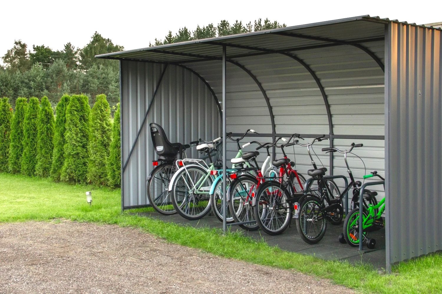 A metal bike shelter with five bicycles and one child's tricycle parked inside, adjacent to a grassy area with green trees in the background.