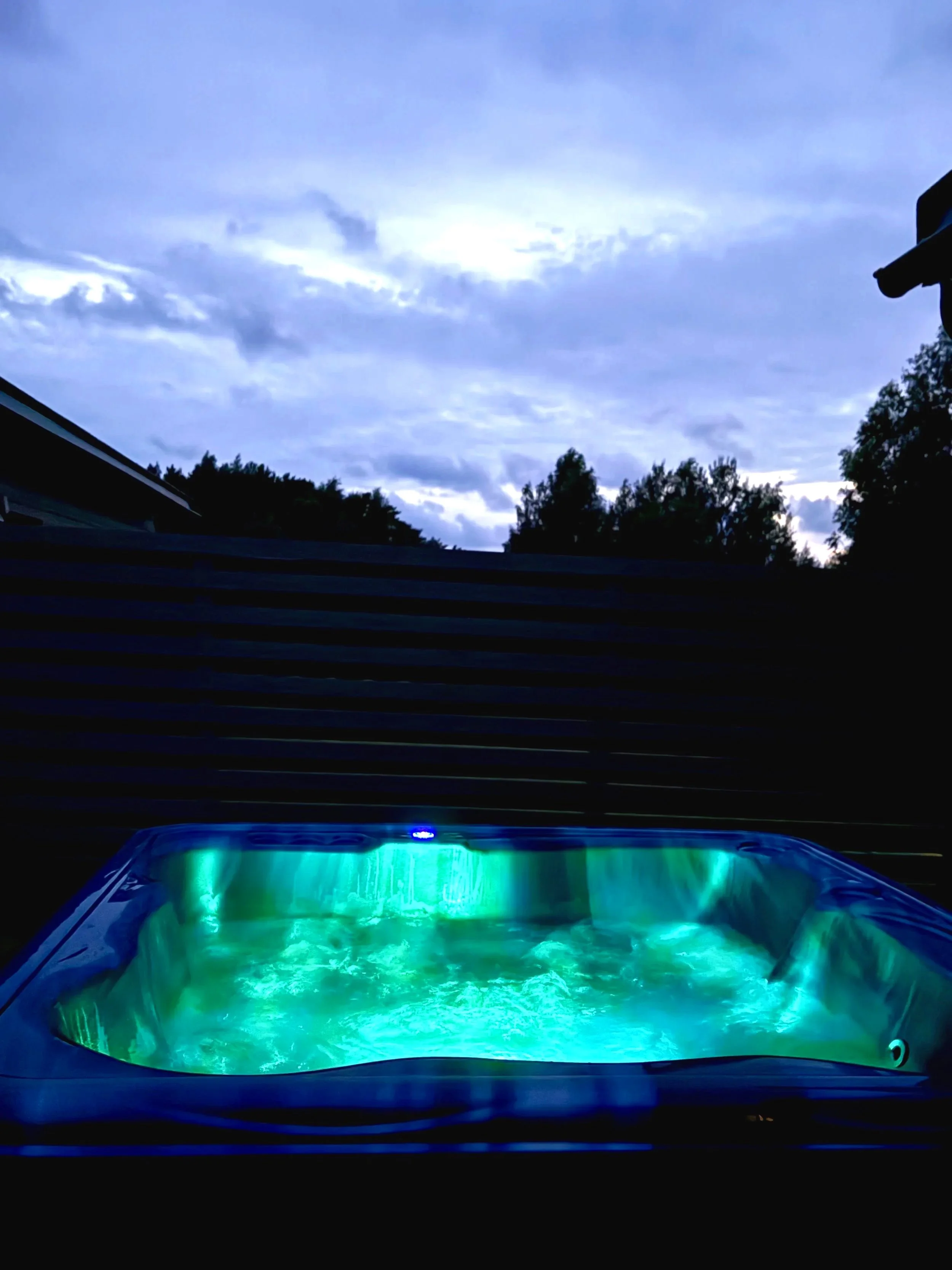 An outdoor hot tub with underwater lights, surrounded by a wooden privacy fence, with a cloudy sky overhead and treetops in the background.