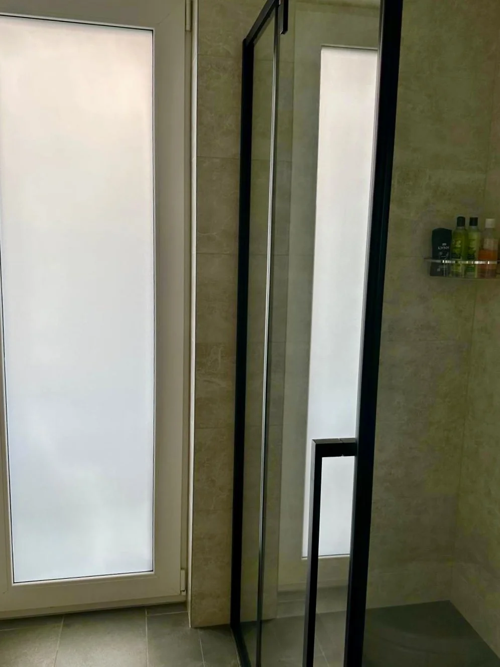 Bathroom with frosted glass door and a walk-in shower with black frame, beige tiled walls, and a shelf with toiletries.