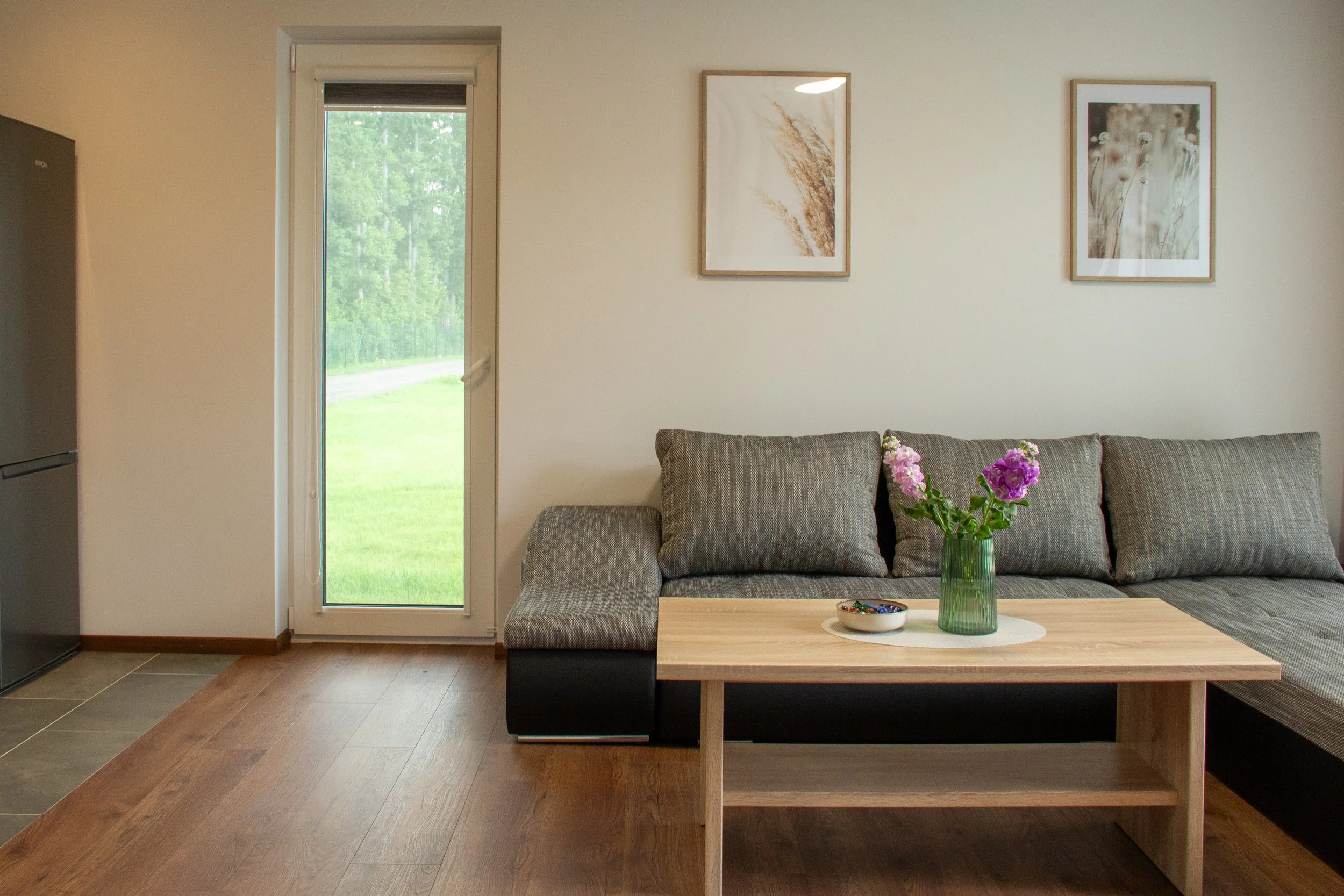 Living room with a beige wall, two framed botanical art prints, a glass door leading outside, a grey sectional sofa with cushions, a wooden coffee table with a vase of pink and purple flowers, and a bowl of candy on a white placemat.
