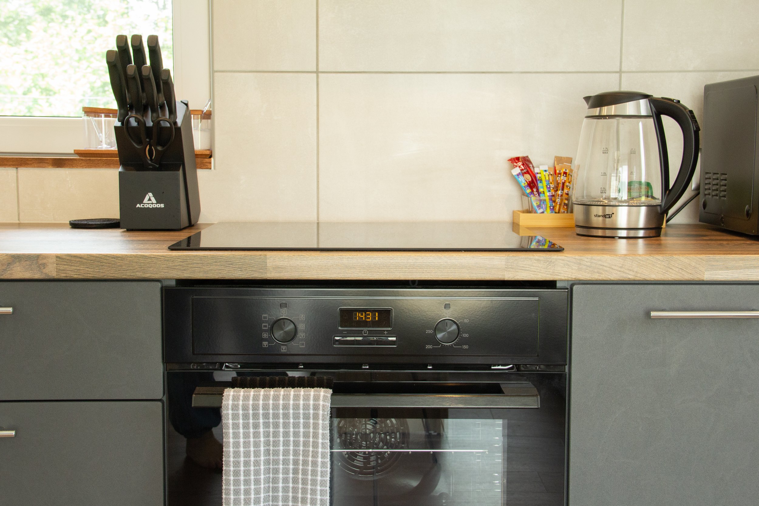Kitchen counter with a black knife block, electric stovetop, electric kettle, and a microwave with a window in the background.