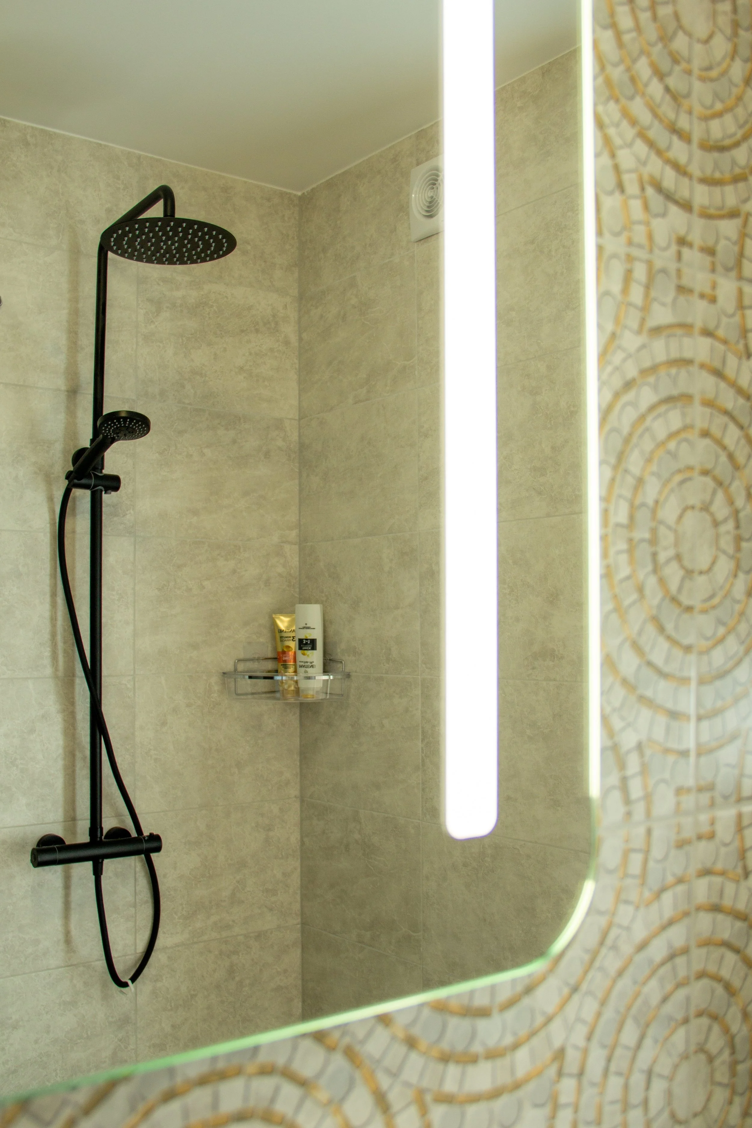 A modern shower area with beige tiled walls, a black showerhead and handheld shower, a niche with toiletries, a vertical mirror with light, and a decorative wall with circular pattern tiles.