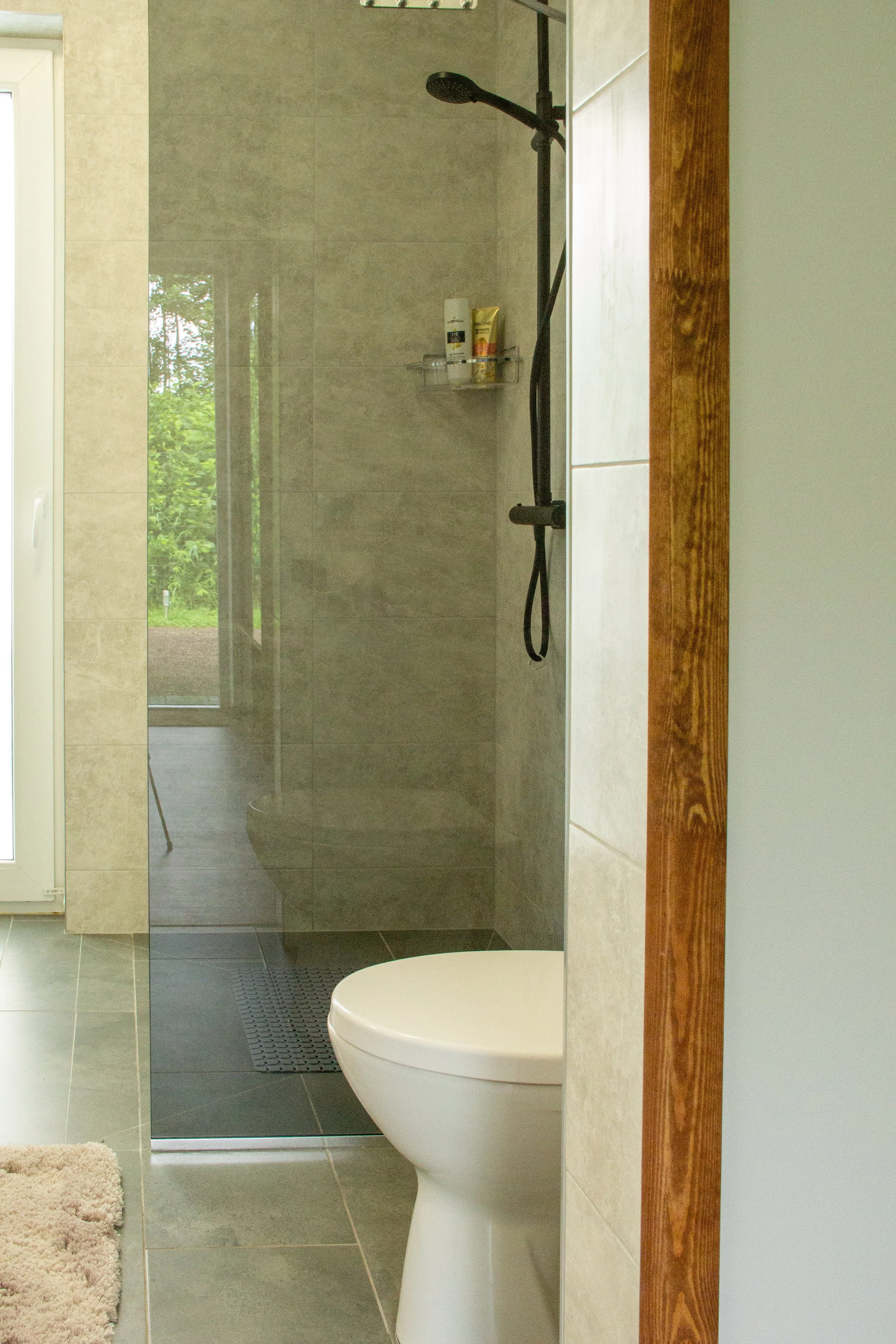 Photo of a modern bathroom with a toilet and a walk-in shower enclosed by a glass partition, a towel on the floor, and a grassy outdoor view through the window.