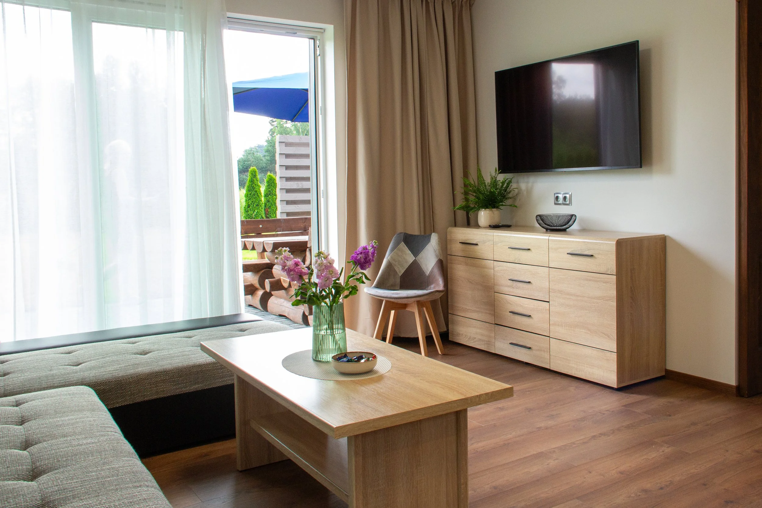 Living room with a sliding door leading to a balcony, wooden flooring, a beige sofa, a wooden coffee table with a vase of pink and purple flowers, a wooden sideboard with a potted plant and a bowl, a wall-mounted flat-screen TV, a chair with a patter