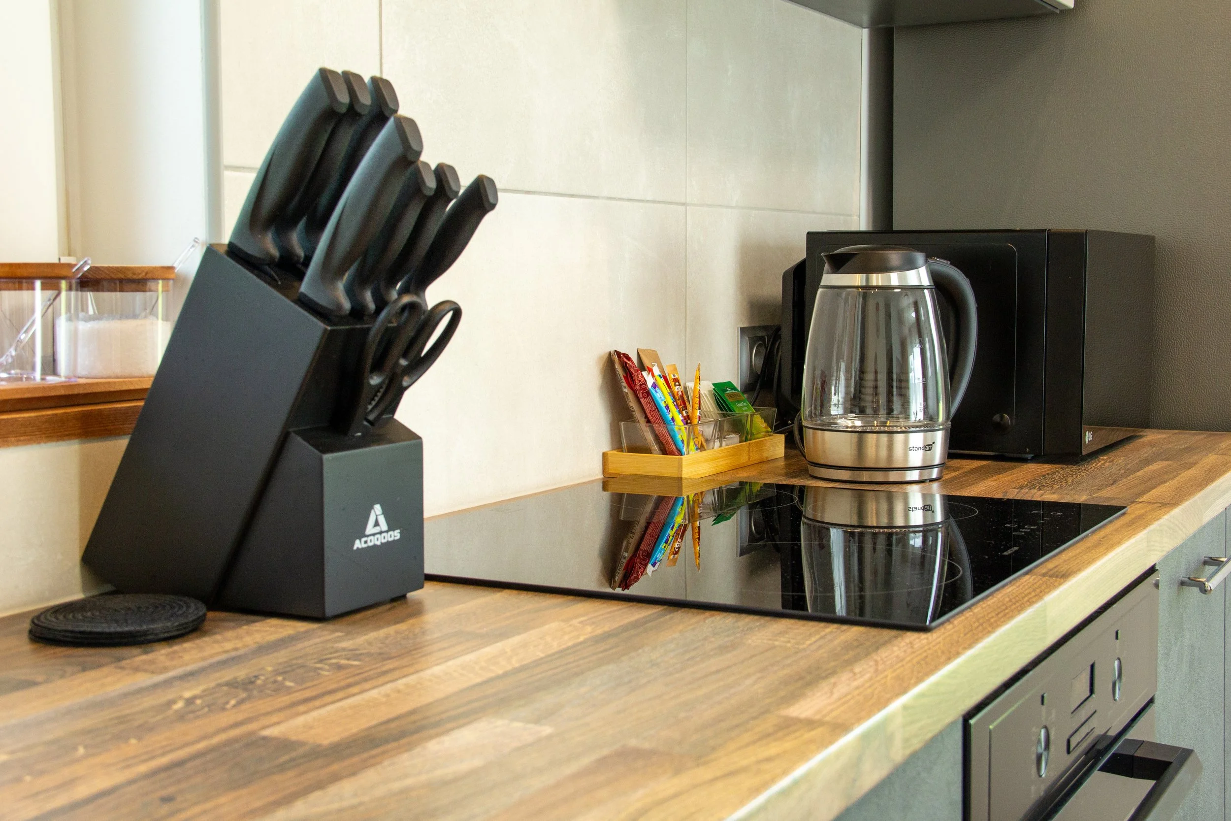 Kitchen countertop with a knife set, a box of tea bags and snacks, a glass electric kettle, and a microwave on a wooden surface.