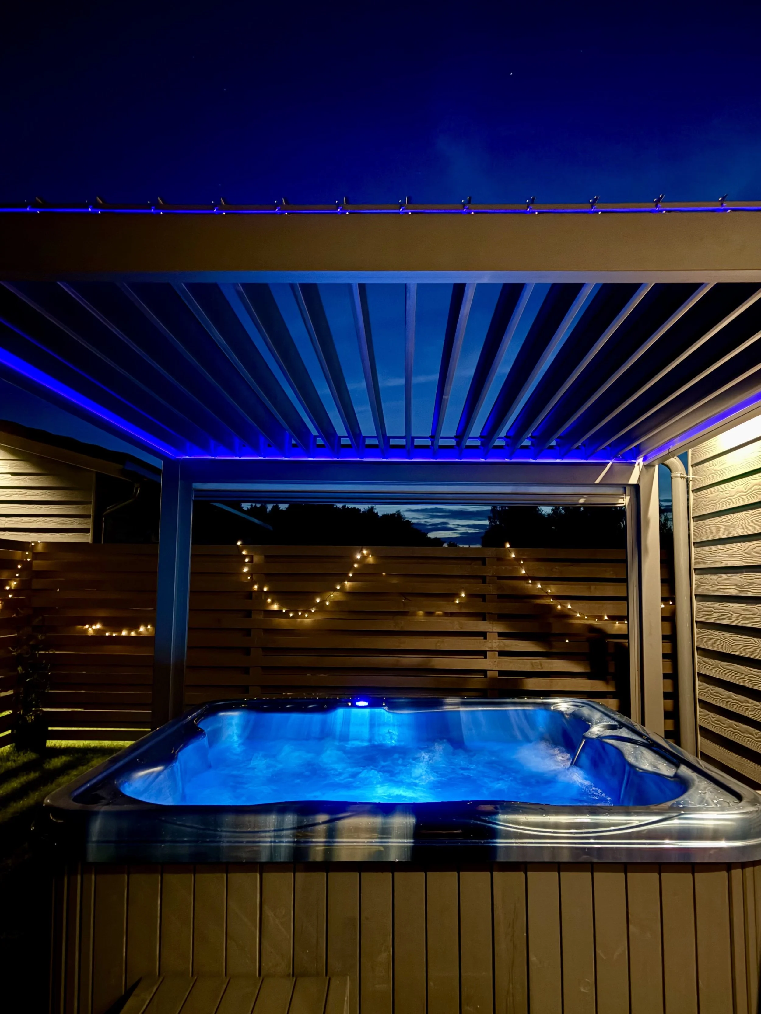 An outdoor hot tub with underwater lights, surrounded by a wooden privacy fence, with a cloudy sky overhead and treetops in the background.