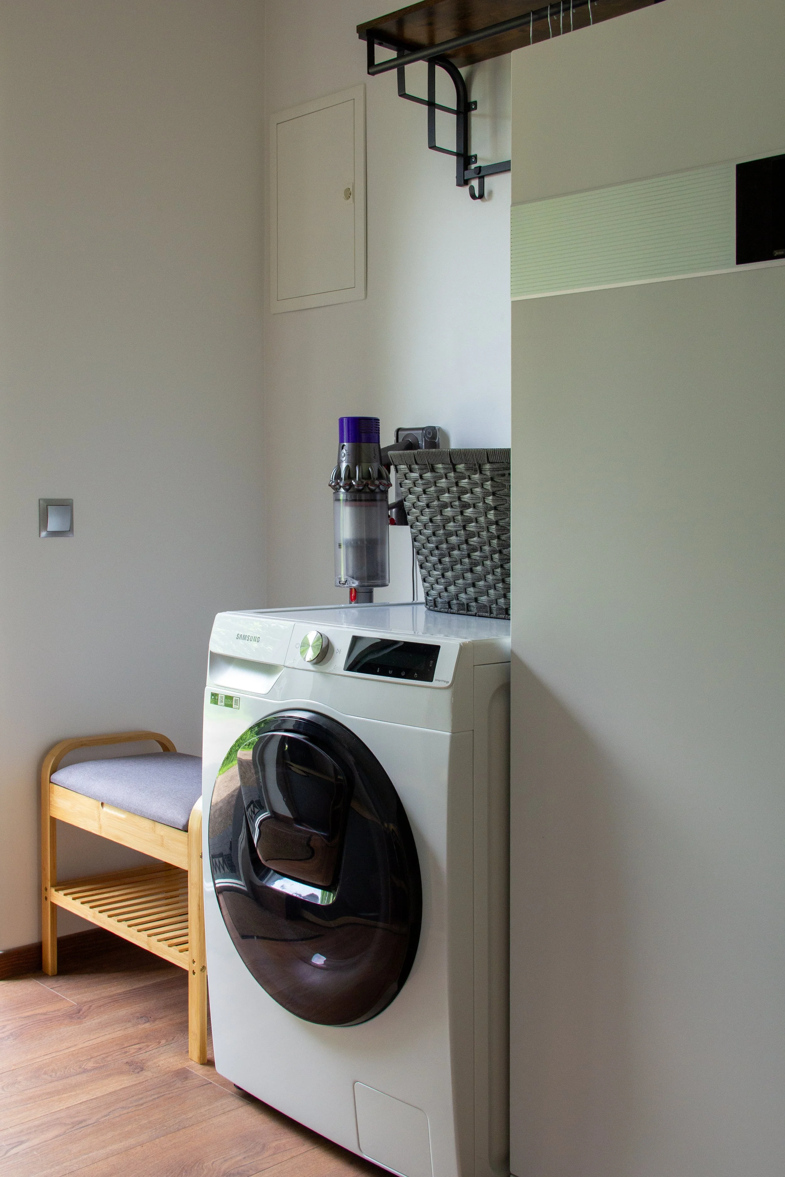 A laundry room featuring a front-loading washing machine, a wooden bench with a cushioned seat, and a basket on top of the machine, with a staircase overhead.