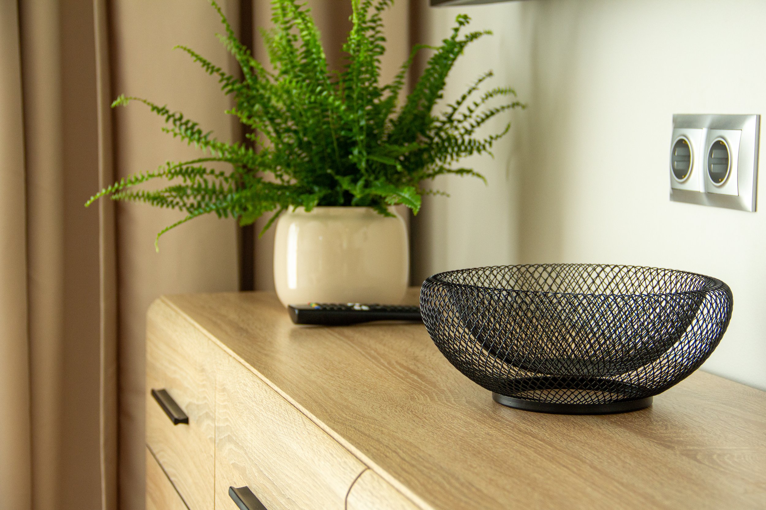 Decorative room interior with a wooden sideboard, a white ceramic vase with a green fern, a remote control, and a black wire fruit bowl.