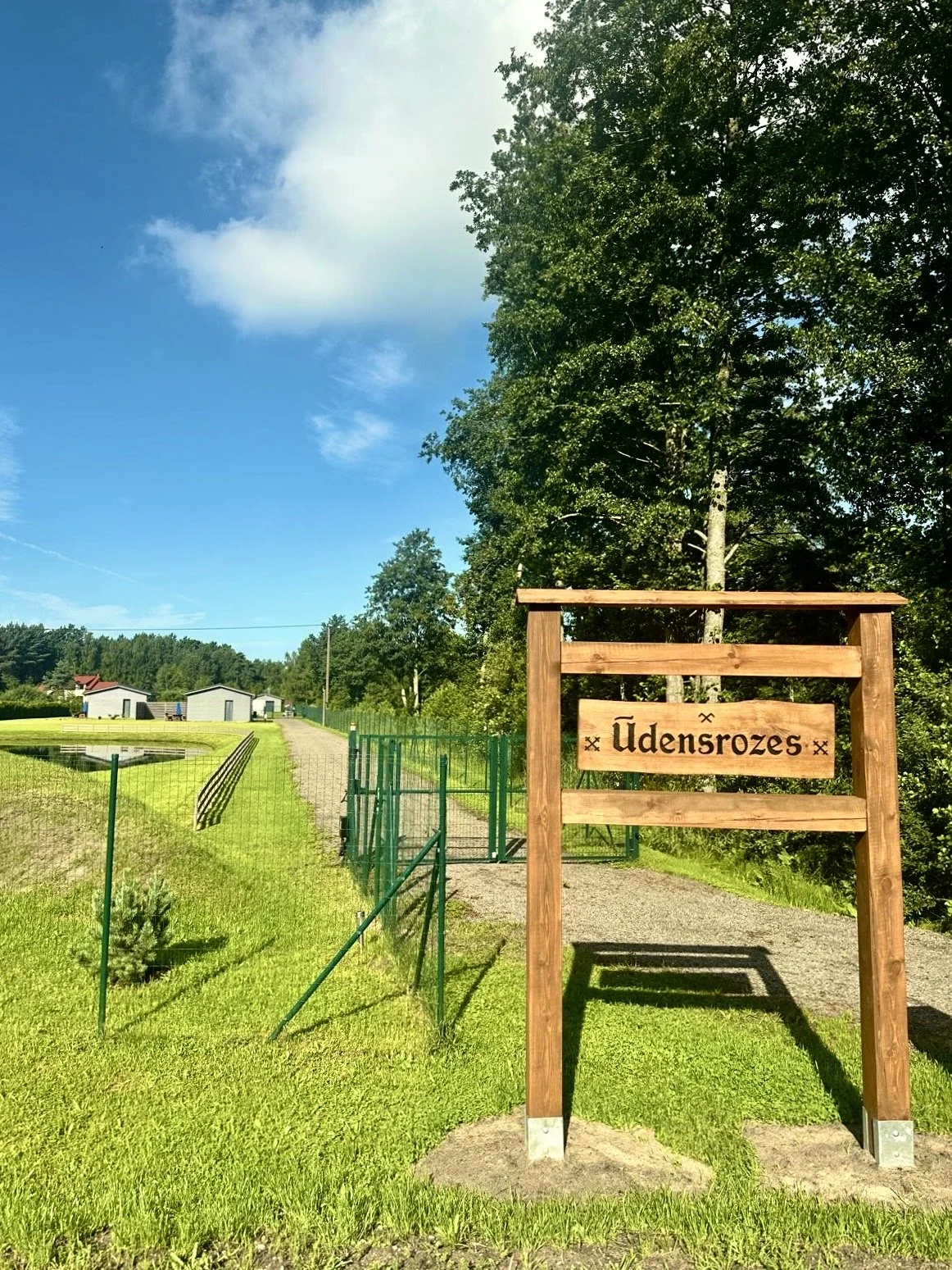 A gravel path next to a green fence running through a rural area with trees, grass, and small buildings in the background. A wooden sign with the word 'Udensrozes' is displayed in the foreground.