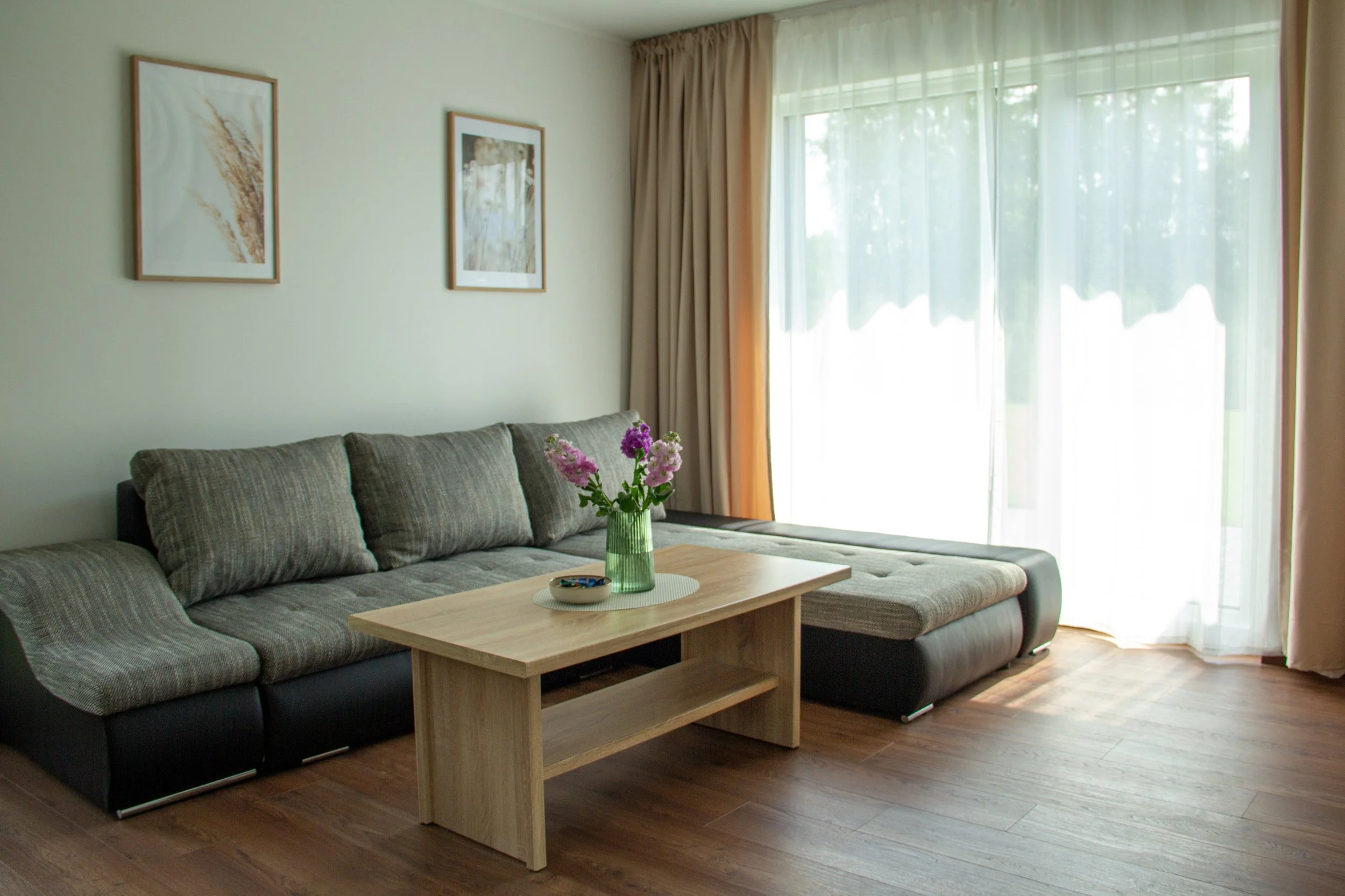 Living room with a large grey sectional sofa, a wooden coffee table with a green vase of pink and purple flowers, curtains, and framed art on the wall.