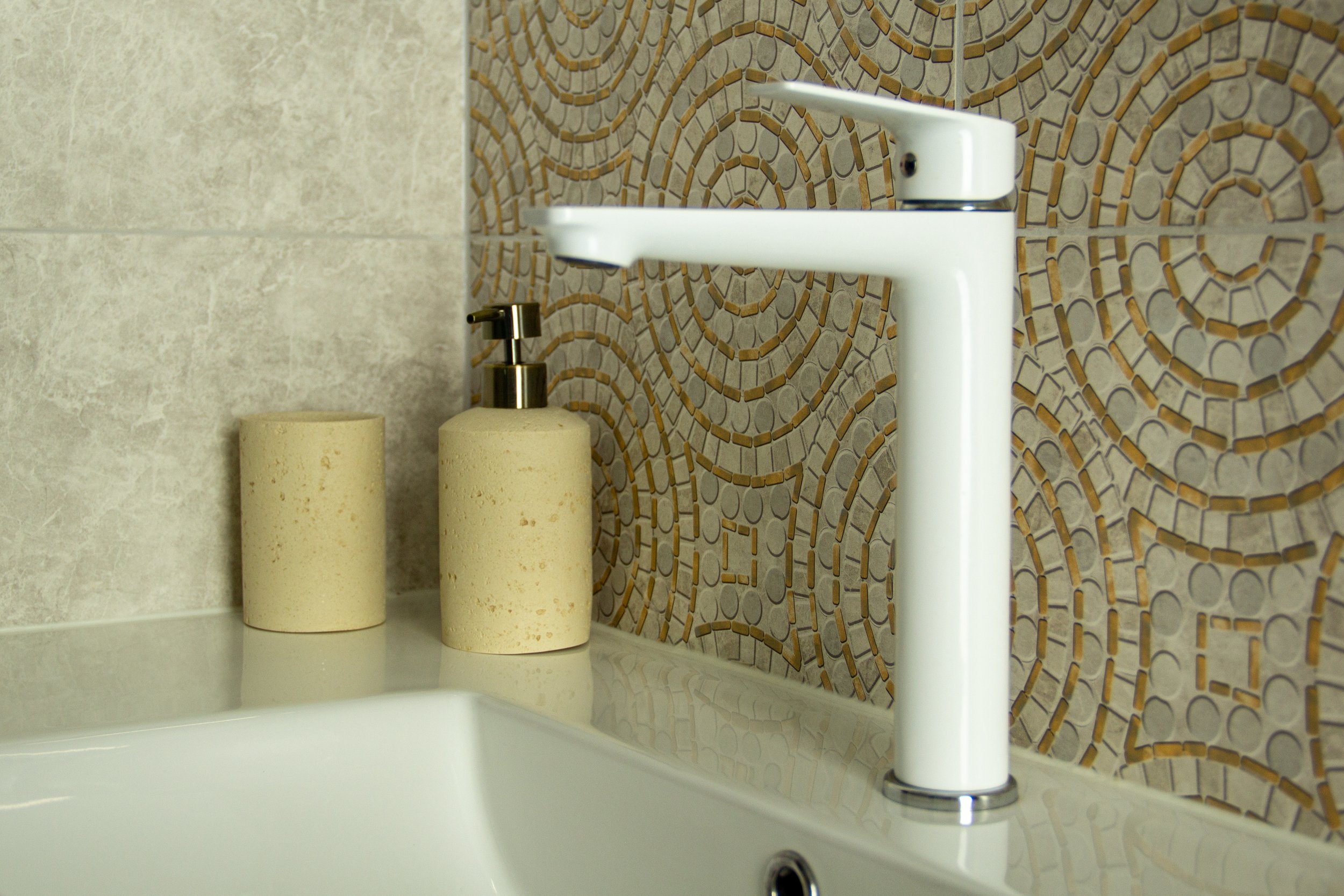 Bathroom sink with white modern faucet, beige soap dispenser, and beige soap dish on a beige countertop. Decorative wall tiles with circular patterns in brown and beige in the background.
